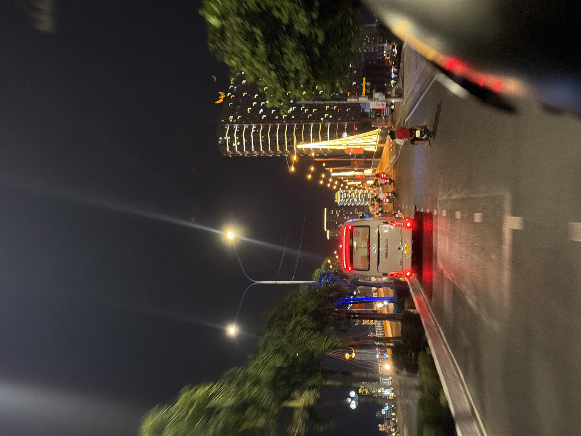 Night scene on a road in Bac Nha Trang Ward, Vietnam. The street is illuminated by street lights. A white van is visible on the right side of the road, along with other vehicles and people further ahead. Buildings are visible in the background, adorned with lights. Trees line the roadside.