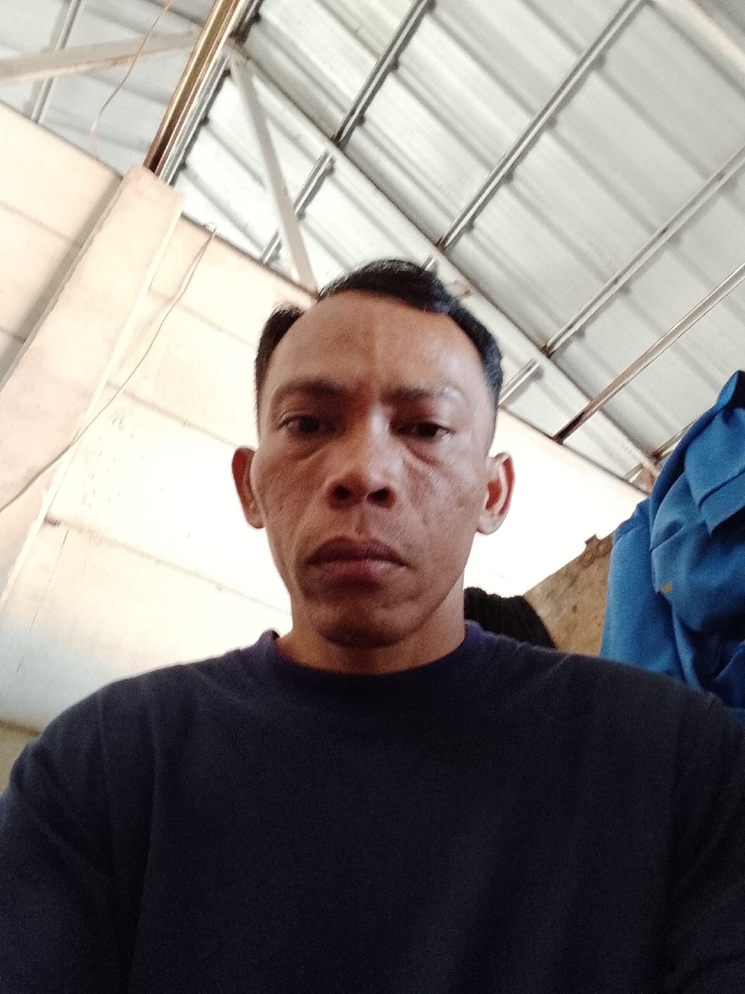 This is a medium shot, point-of-view photograph of a man in Bukit Ketok, Indonesia. The man is looking directly at the camera. He has dark hair, a dark complexion, and is wearing a dark blue shirt. The background consists of a corrugated metal roof with visible supports, and a light-colored wall. A blue garment is visible on the right side of the frame. The lighting suggests an indoor or covered outdoor environment, possibly a workshop or storage area.
