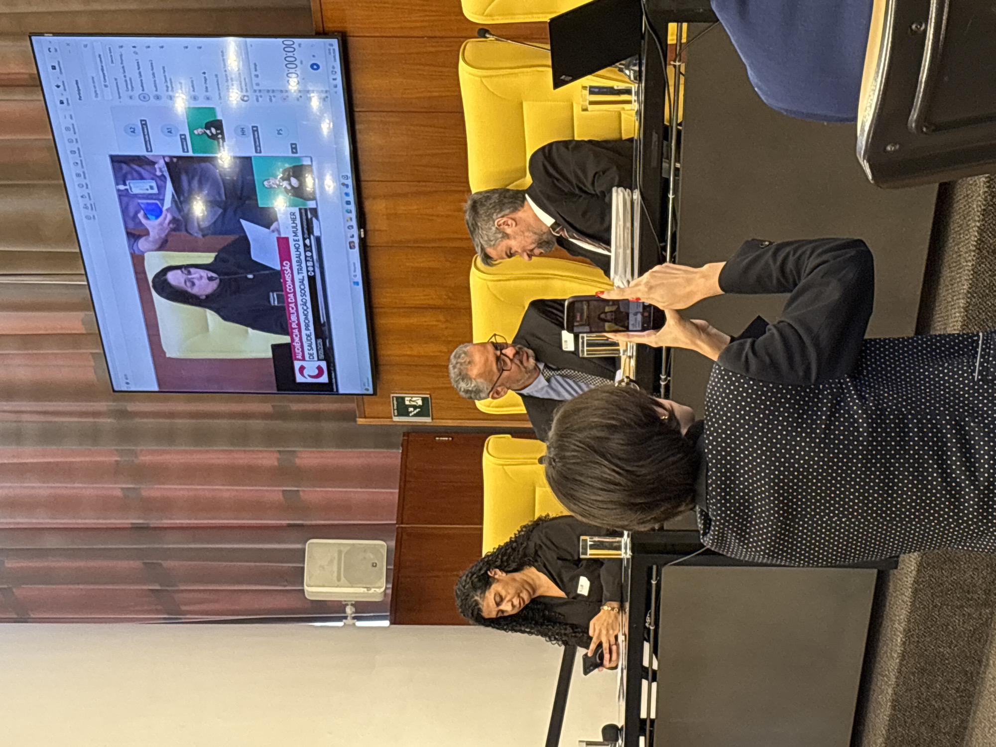 The image shows a group of people attending a public hearing in a room. The room appears to be an auditorium or conference hall with yellow upholstered seats. There is a large screen displaying a video conference or live stream of a woman speaking. Text on the screen reads "AUDIÊNCIA PÚBLICA DA COMISSÃO DE SAÚDE, PROMOÇÃO SOCIAL, TRABALHO E MULHER," which translates to "Public Hearing of the Commission on Health, Social Promotion, Labor and Women." This implies the hearing is related to health, social issues, labor, and women's affairs. Several people are seated at a table, including two men in suits and a woman with curly hair. One person is taking a picture of the screen with their phone. A sign with a white arrow pointing to the right is visible on the wall. The exact location in the city of São Paulo, Brazil, is not identifiable from the image alone.
