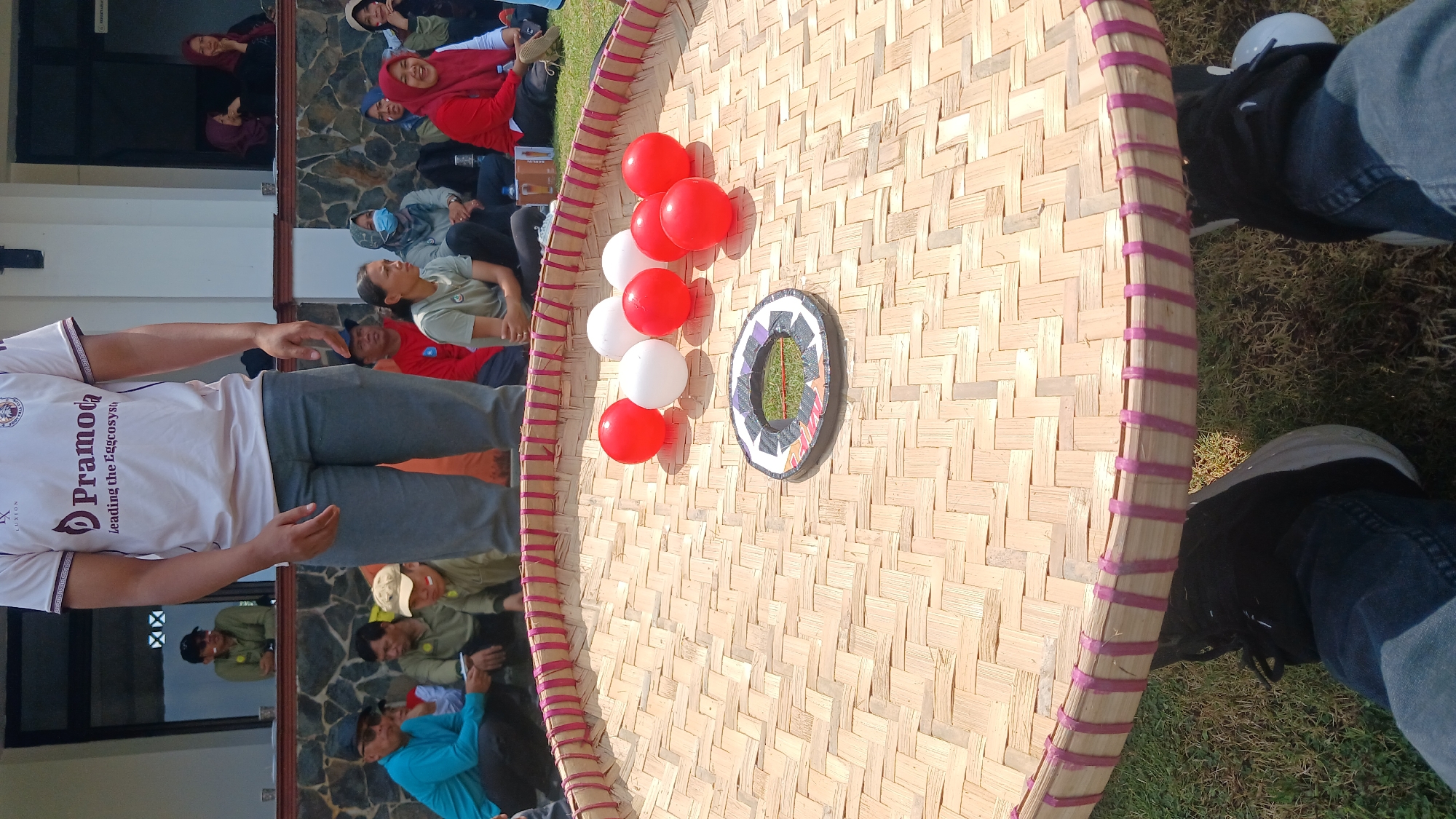 In Ngasinan, Indonesia, a group of people are gathered outdoors for a game. In the foreground, a large, round woven bamboo tray, approximately 1 meter in diameter, is laid out on the grass. The tray has a decorative pink woven edge. On the tray are seven ping pong balls, four red and three white, and a small, circular target with colorful geometric patterns and what appears to be grass in its center. In the midground, a person wearing a white t-shirt with the logo "Pramoda" and grey pants stands, partially obscuring the view of the onlookers seated behind the tray. The onlookers, dressed in casual attire, are watching the game. Some are seated on the grass, while others are positioned on a raised platform behind them. The background shows a building with a white facade and windows. The overall scene suggests a daytime event in an outdoor recreational or community setting.
