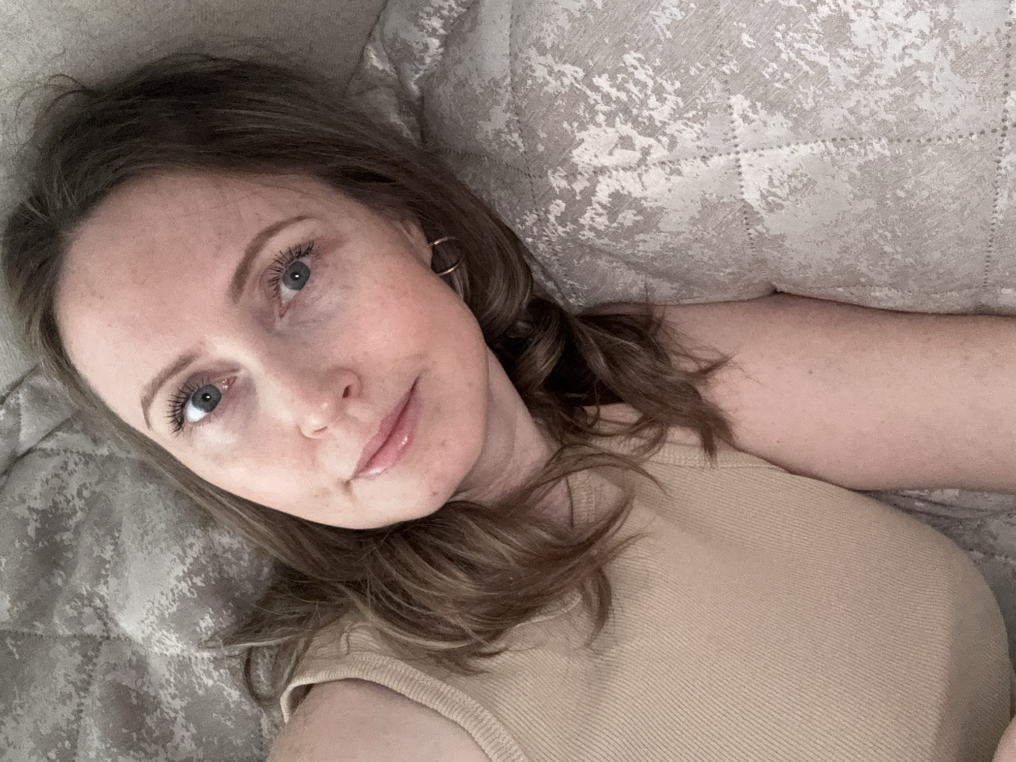 A woman lies on a bed or couch with her head resting on a pillow. She has fair skin, light brown hair, and blue eyes. She is wearing a light beige, sleeveless top. She is wearing small hoop earrings. The background consists of a textured, light gray and white pillow. The image does not provide sufficient information to determine its location within the city of Chaikovsky, Russia, or confirm that it is located there at all.