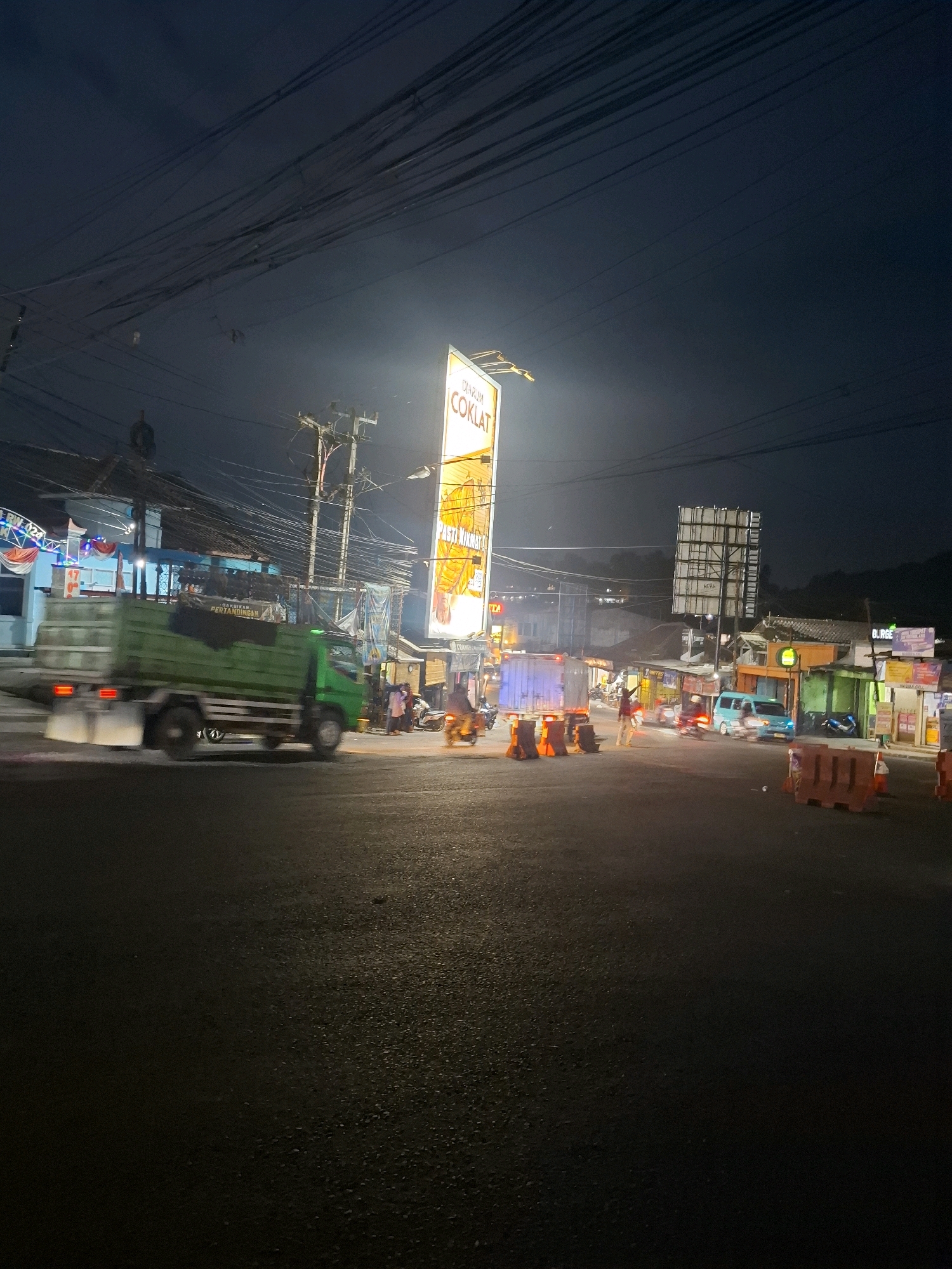 This image depicts a street scene in Cibadak, Indonesia, at night. The road is asphalt, and several vehicles are visible, including a green truck in motion, a light blue van, and several motorcycles. There are also temporary orange traffic barriers on the road. Buildings line the street, some illuminated by signs and lights. A tall billboard advertising "DIARUM COKLAT" (Djarum Chocolate) is prominently displayed. Power lines are strung overhead, characteristic of many urban areas. Several individuals are present, some walking and others on motorcycles, suggesting ongoing activity despite the late hour. The overall lighting is dim, with light sources from streetlights, vehicles, and signs casting illumination on the scene.