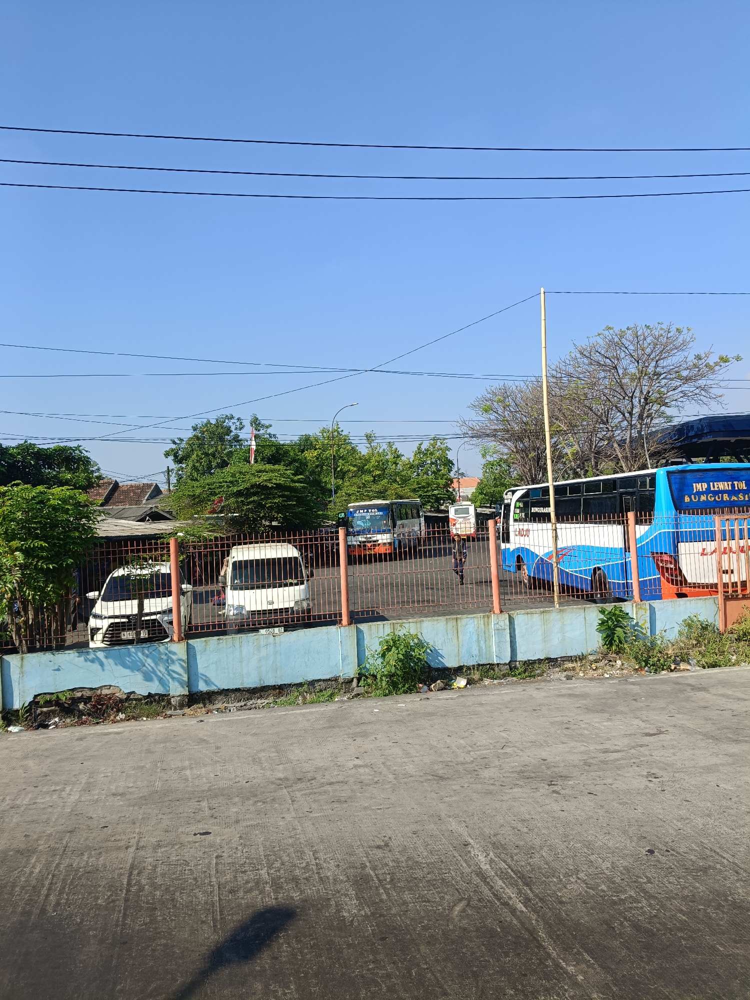 The image shows a street scene in Kedungrejo, Indonesia. In the foreground, there is a paved area with tire marks and a shadow of a lamppost. Behind this, a fence separates the paved area from a lot containing several vehicles, including two white cars and multiple buses. The buses are parked in what appears to be a bus terminal or depot. One bus, predominantly blue and white, has "JMP TOL LEWAT TOL" and "BUNGURASI" written on its side. Another bus, further to the left, is mostly white with blue accents. In the background, there are trees, a building with a red tile roof, and a clear blue sky with power lines. The overall impression is a sunny day at a transportation hub.