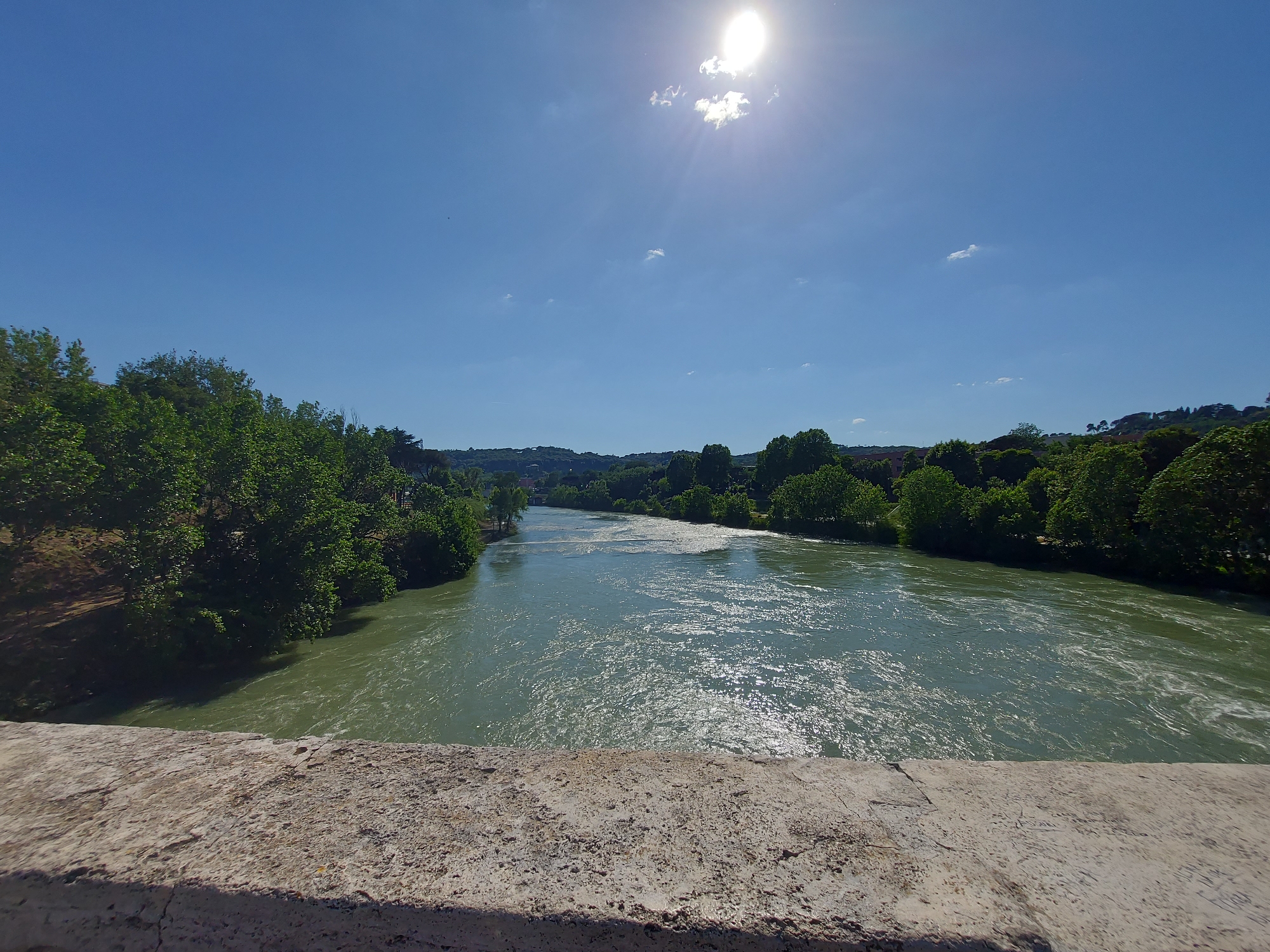 The image depicts a view of the Tiber River in Rome, Italy, on a sunny day. The perspective is from a bridge, showing a textured concrete barrier in the foreground. The river flows from the distance towards the viewer, its surface reflecting sunlight with visible currents and ripples. The riverbanks are lined with lush green trees and vegetation. The sky is a clear blue, with the sun positioned high in the frame, partially obscured by a small cloud.