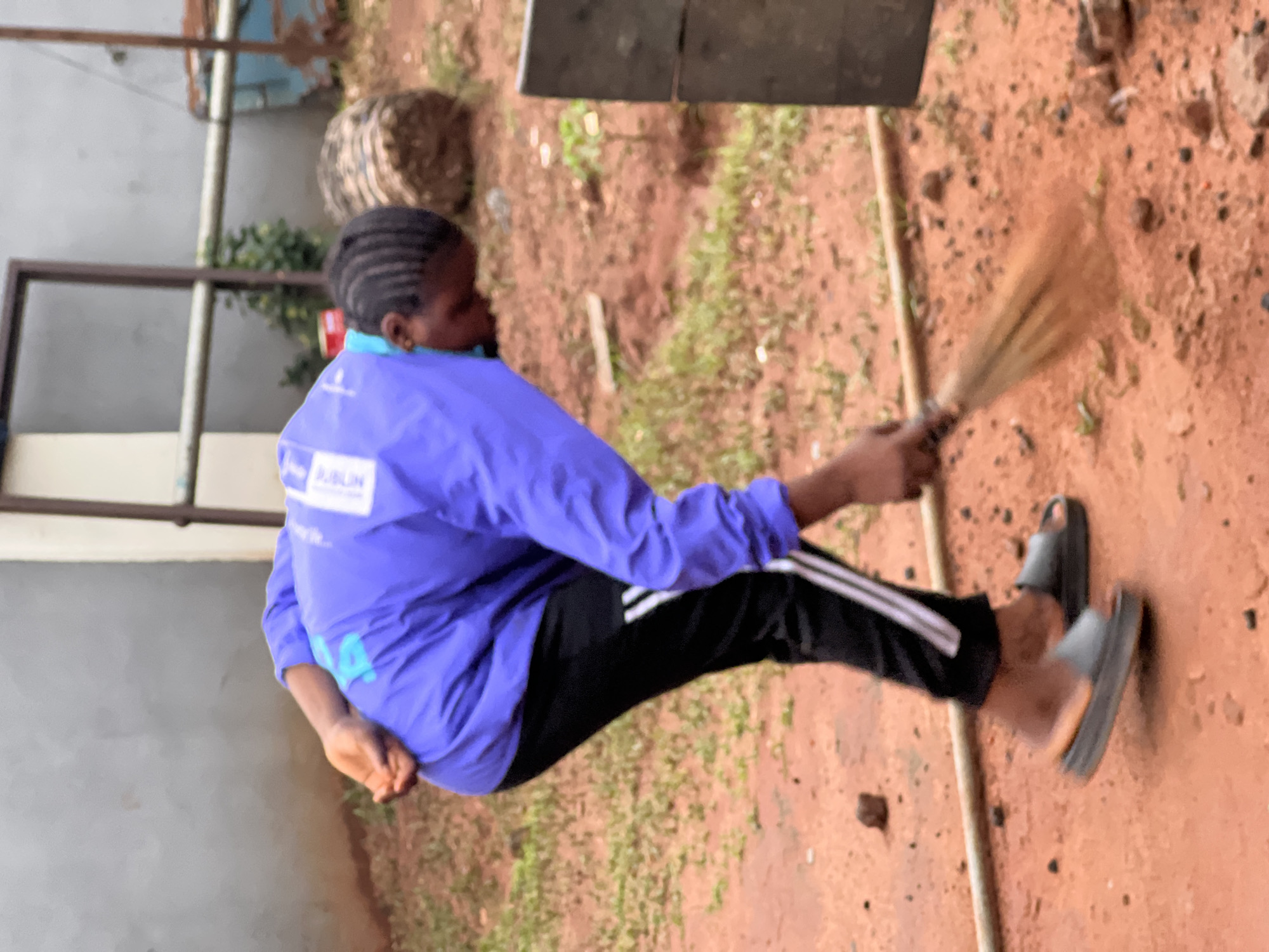 In the city of Nkpor, Nigeria, a person is captured in a medium shot, bent over and sweeping a patch of reddish-brown dirt ground with a broom. The individual, with braided dark hair, wears a bright purple athletic jacket with white accents on the sleeves and black track pants with white stripes down the sides. They are also wearing dark gray slides. The background features a textured gray wall, a metal frame structure, and some vegetation. The overall environment appears to be an outdoor, somewhat unkempt area, possibly a yard or communal space. The focus is on the action of sweeping.