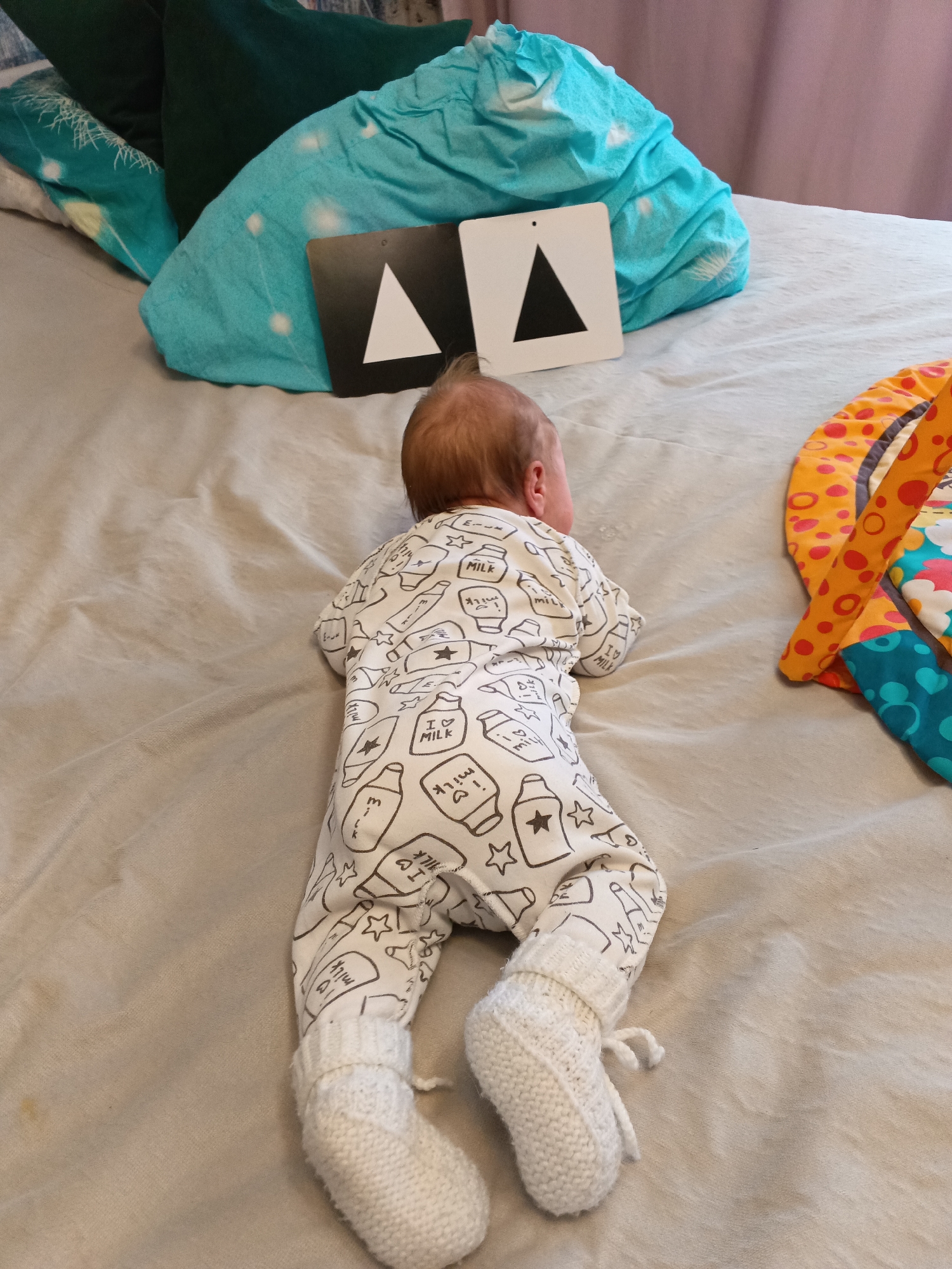 A baby in a milk-bottle-patterned onesie and knitted white socks lies face down on a bed. Two black-and-white cards with triangle patterns are positioned near the baby's head. Green and turquoise pillows are behind the cards. A multicolored play mat is to the right of the baby. The scene is set in what appears to be a private residence. No information is present to suggest the location is in Andreyevo, Russia.