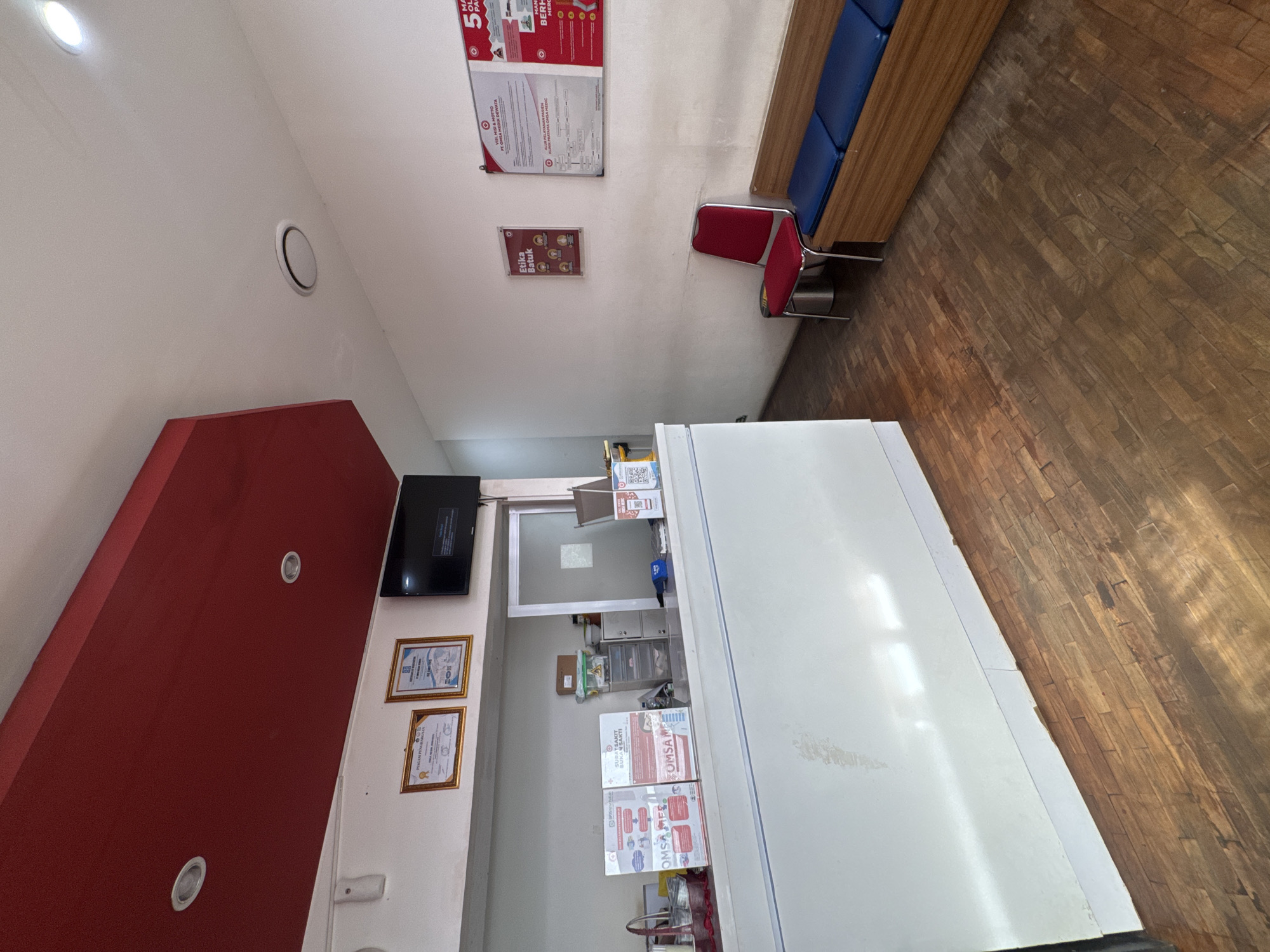 The scene appears to be an interior shot of a clinic or medical office in Kuta Selatan, Indonesia. There is a white reception desk, a red accent wall with recessed lighting, and a television mounted on the wall. Framed certificates are hung near the TV. Informational posters in Indonesian are visible on the walls, including one about "Etika Batuk" (cough etiquette) and another with the acronym "OMSA." A waiting area with blue cushioned seats and a red chair is also present. The floor is made of wood planks. A partial view of a doorway suggests an inner room with shelves.