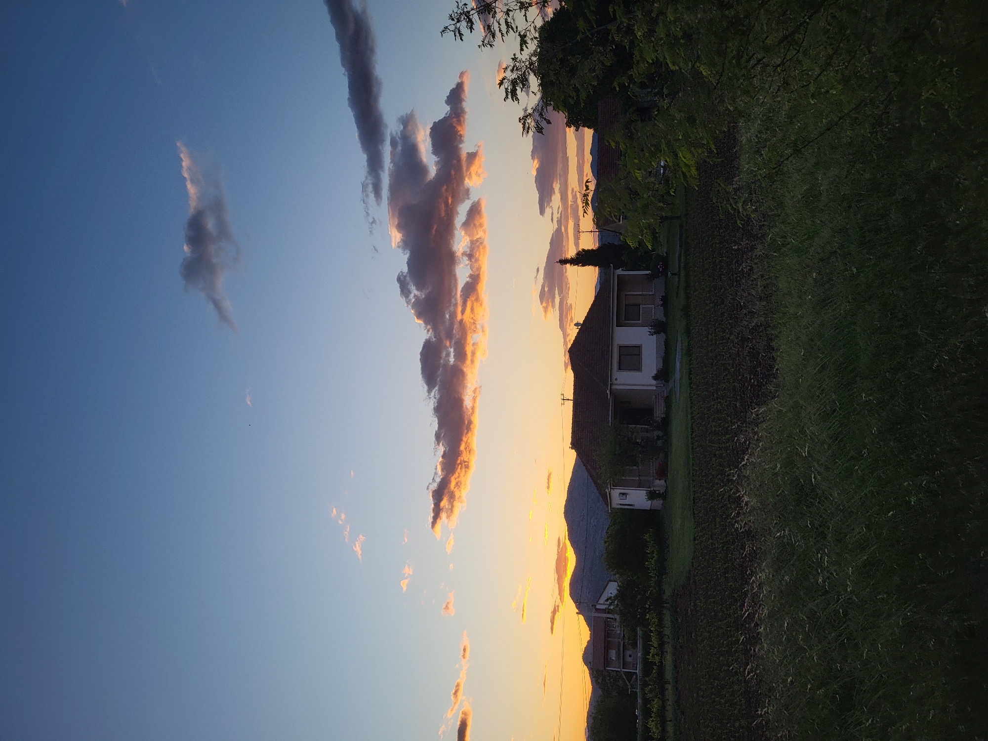 The image captures a serene rural landscape at what appears to be sunset. The sky, dominating the upper two-thirds of the frame, displays a gradient of colors, transitioning from a clear, deep blue in the upper left to vibrant oranges, yellows, and warm pinks closer to the horizon on the right. Wispy, scattered clouds are visible, with some darker formations against the blue sky and others brilliantly illuminated from below by the setting sun, appearing golden and fiery. A prominent, elongated cloud formation stretches horizontally across the sky, catching the most intense light.

In the lower third of the image, the landscape unfolds. Several houses with dark roofs and light-colored walls are nestled within the scene, partially silhouetted against the bright horizon. These structures suggest a small village or hamlet. Behind the houses, the dark silhouette of a mountain range is visible against the glowing sky. The foreground and midground feature lush green vegetation, including fields of grass and scattered trees with dense, dark foliage. The lighting suggests the sun is very low on the horizon, casting long shadows and bathing the clouds in warm light. No individuals or specific activities are depicted. There is no visible text. The scene evokes a tranquil and peaceful atmosphere, characteristic of a late evening in a natural setting, consistent with the location of Mahala, Montenegro.