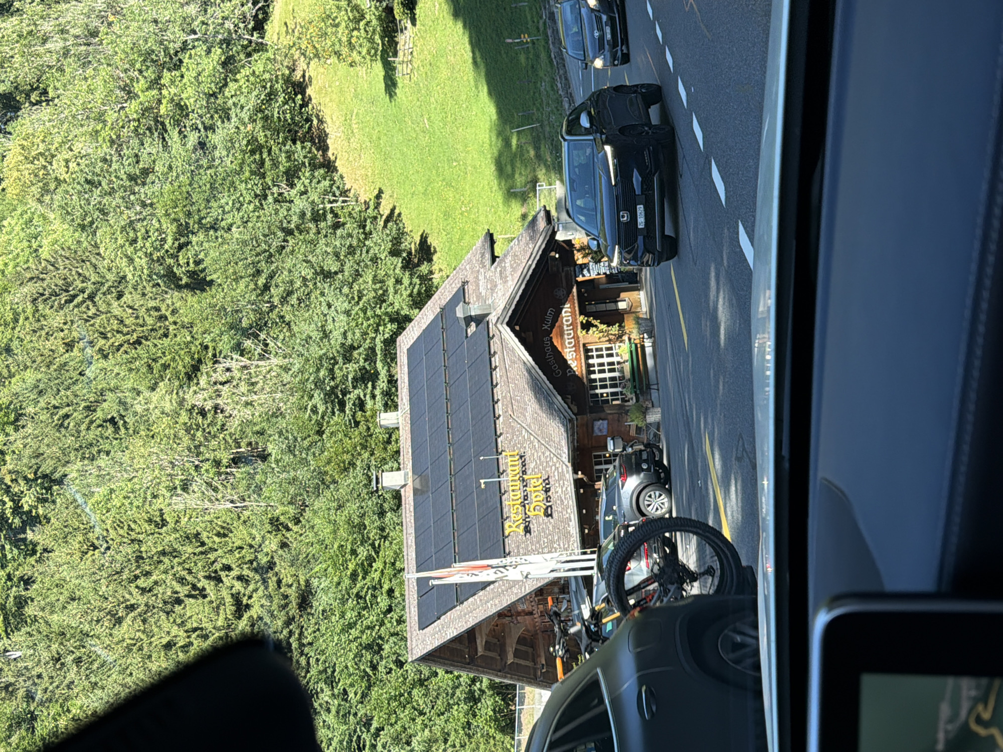 The image shows a street scene in a mountainous, forested area, likely in Europe. In the foreground, a dark-colored car is visible from the interior, with a dashboard and part of a screen showing a map. To the left of the car, a dark SUV with a bicycle mounted on its rear is parked. Further down the street, a black Honda SUV is driving, with a Swiss license plate reading "ZF-10629". To its right, another dark-colored car is also driving.
Dominating the mid-ground is a rustic, wooden building with a pitched roof covered in solar panels. The building appears to be a restaurant or hotel, with signage that reads "Restaurant Hotel" in yellow lettering and "Gasthaus Kulm Restaurant" on the lower part. There are flags outside the building. The area around the building is paved, with a yellow line marking the road.
The background features a steep, grassy slope on the right, dotted with trees and a wooden fence. A dense forest of evergreen trees covers the hillside on the left. The overall impression is a scenic, rural location, possibly a mountain pass or village. The lighting suggests it is daytime with clear skies.