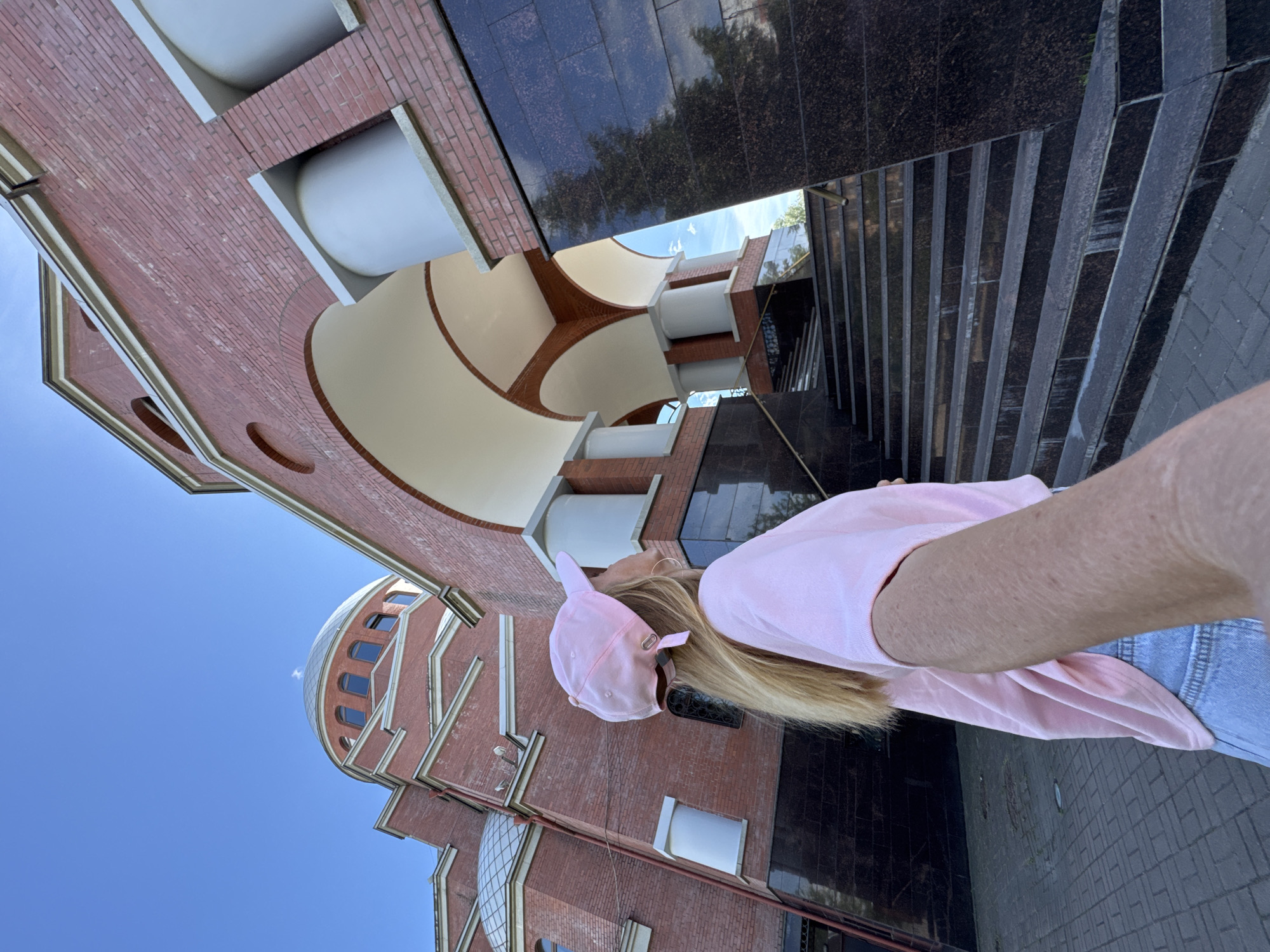 A woman with blonde hair wears a pink t-shirt and a pink baseball cap. She is positioned in the foreground of the image, with her right arm extending upwards and to the right, suggesting she is taking a selfie. The background features a red brick building with a domed section and arched architectural elements. The building's exterior is adorned with decorative white and tan molding. Adjacent to the building are dark granite steps and a sidewalk paved with grey bricks. The sky is a clear, bright blue, indicating a sunny day. The orientation of the image is skewed, with the building appearing to stretch upwards and the ground beneath it. There is no visible text in the image.