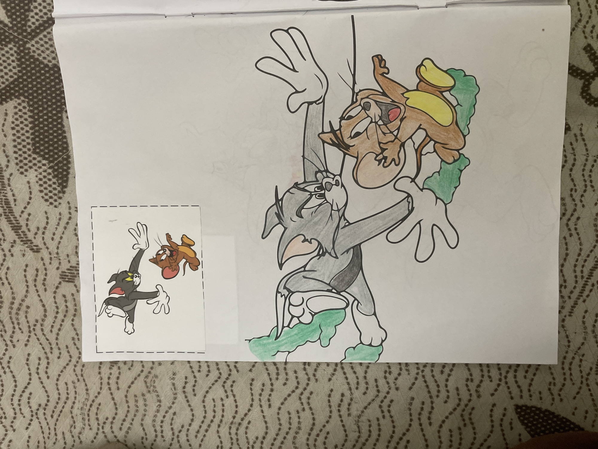A colored drawing of the animated characters Tom and Jerry is featured on a sheet of white paper. The main drawing depicts Tom, the cat, in shades of grey and white, looking upwards with a dynamic expression, his body positioned as if climbing or falling. He is entangled with bright green, leaf-like foliage. Jerry, the brown and tan mouse, is positioned above and to the right of Tom, clinging to the same green foliage with yellow paws, his mouth open in an active expression, seemingly interacting with Tom.
To the bottom left of the main drawing, there is a smaller, separate paper cutout or sticker, framed by a dashed line border. This cutout also shows Tom and Jerry in a different, energetic pose, appearing to be mid-air, possibly falling or in a chase. Tom in this cutout is darker grey/black and white, and Jerry is brown and orange.
The entire paper with the drawings rests on a textured, patterned fabric surface, likely a blanket or bedspread, in shades of beige and brown with wavy line patterns. The artwork is colored with what appears to be crayons or colored pencils. There is no visible text other than the dashed border around the small cutout.
