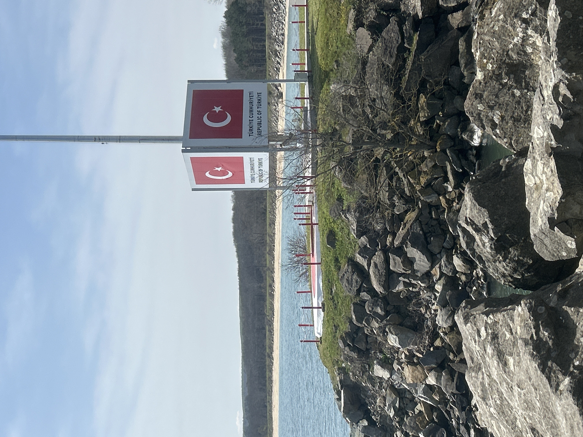 The image shows a border crossing into Turkey. The image includes a sign that states "Türkiye Cumhuriyeti, Republic of Türkiye" with the Turkish flag. The location appears to be near the city of Rezovo, Bulgaria. The border is marked by red and white posts, and the landscape includes water, a sandy area, trees, and rocky terrain. No people are visible.