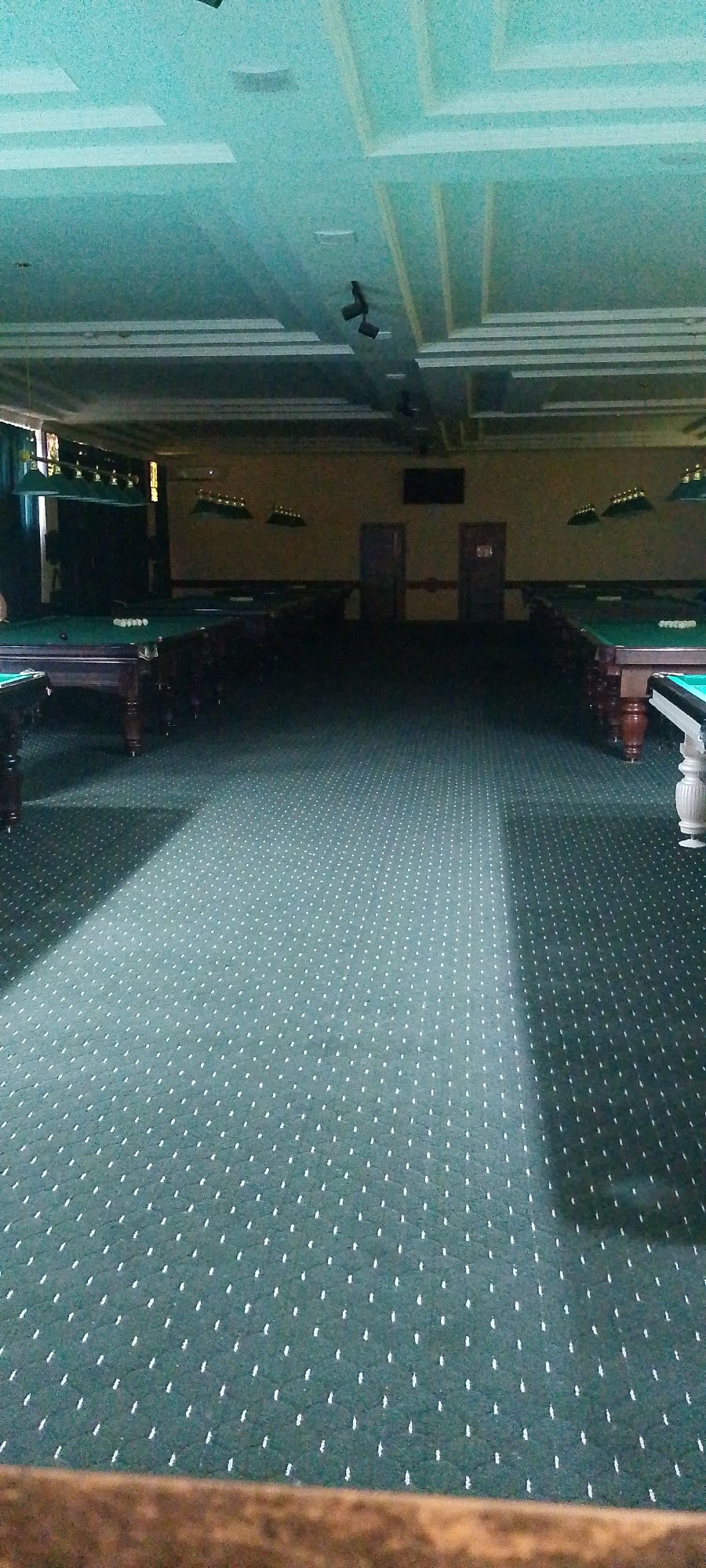 The image shows a dimly lit indoor billiards hall with several pool tables arranged in rows. The room has a teal-colored ceiling with a tiered design and recessed lighting fixtures. The walls are a light beige color, and there are two doorways visible in the background. The floor is covered with a dark green carpet featuring a repeating white dot pattern. Pool tables, each with green felt surfaces and visible balls, are the primary objects in the scene. Several green lampshades are suspended above the tables, providing illumination. The overall atmosphere is that of a functional, but somewhat dated, entertainment venue. The location is identified as a billiards hall in Angren, Uzbekistan.