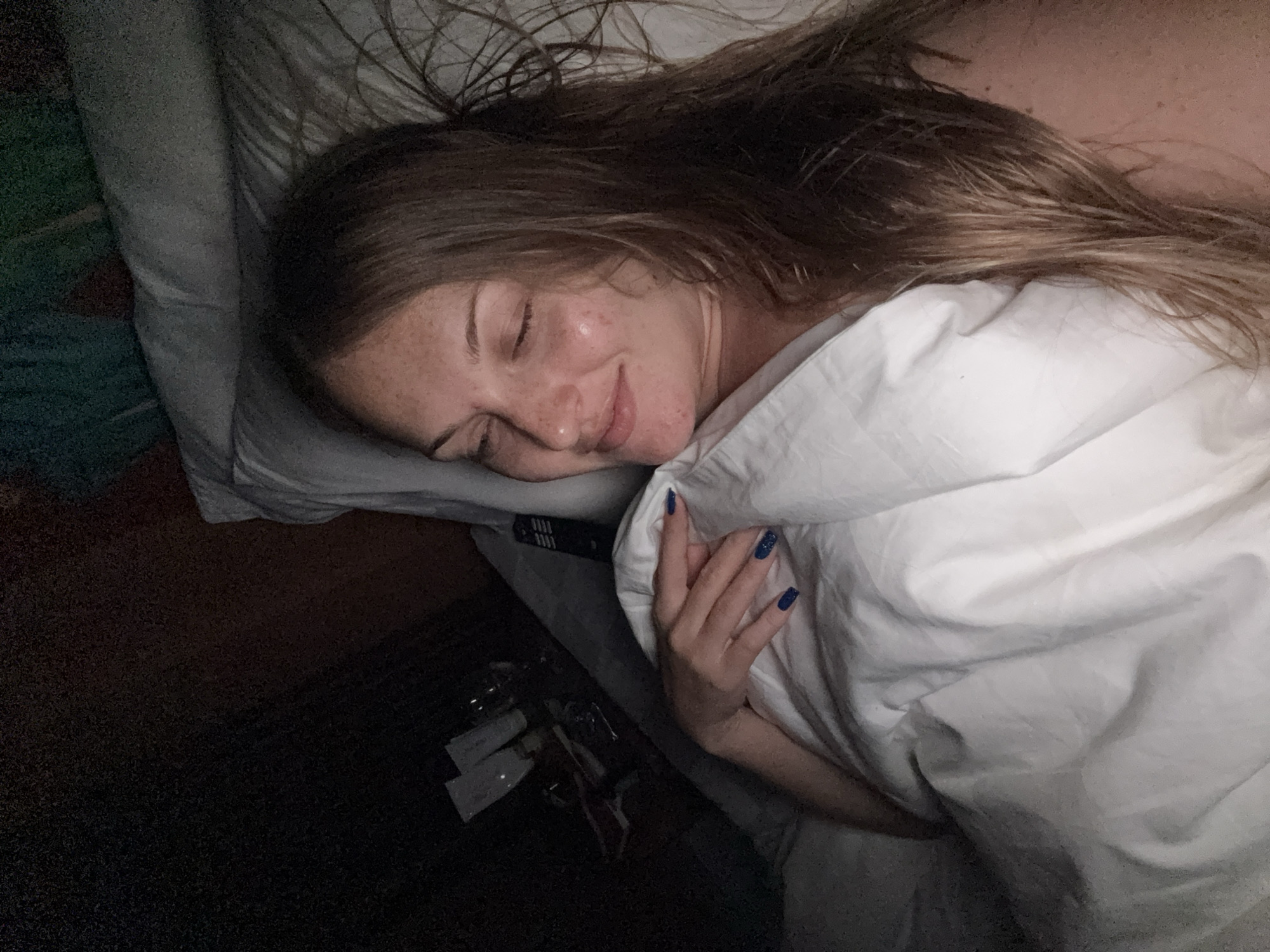 In a dimly lit bedroom in Denpasar, Indonesia, a young woman with long brown hair and freckled skin lies in bed, eyes closed and a gentle smile on her face. She is partially covered by a white duvet. Her left hand rests on the duvet, revealing dark blue nail polish. To her left, on a bedside table, are several bottles, likely toiletries or cosmetics, and a black remote control. The overall atmosphere is one of peaceful slumber in a domestic setting.