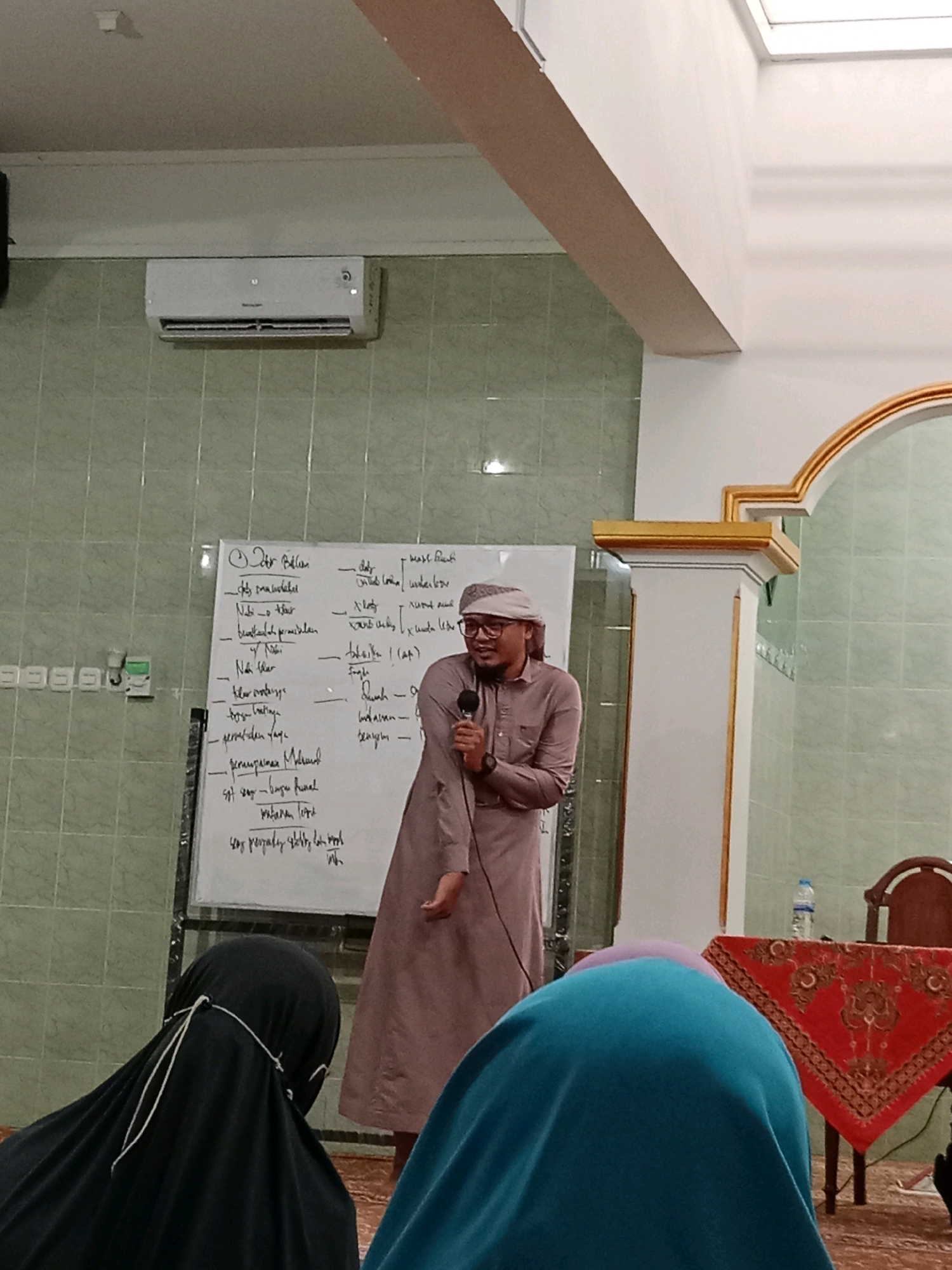 A man wearing a light brown robe and a white keffiyeh, glasses, and a microphone is standing and speaking in front of a whiteboard in what appears to be a classroom or community hall. The whiteboard has writing on it, some of which is legible. The man is smiling and gesturing with his left hand. In the foreground, two people are seated, their backs to the camera, one wearing a black hijab and the other a teal hijab. To the right, a table is set with a red tablecloth, a bottle of water, and a chair. The room has tiled green walls and a white ceiling with an air conditioning unit. The architectural elements suggest a possible mosque or Islamic center. The location is the city of Cemani, Indonesia.