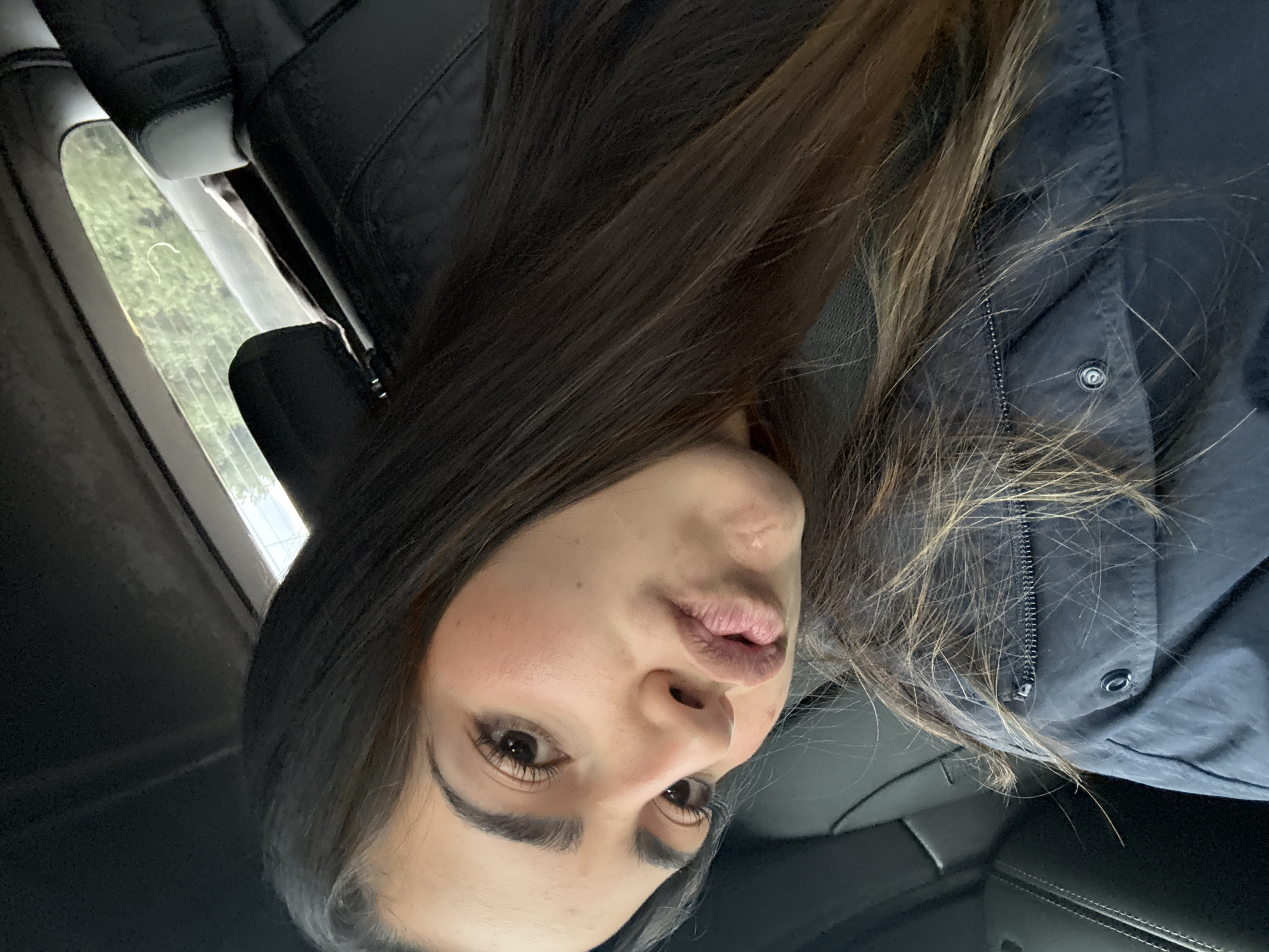A person with long, dark hair is sitting inside a vehicle. The individual is wearing a dark jacket with a visible zipper and button. The scene includes a view out the car window of what appears to be trees or foliage. The interior of the car includes a headrest and possibly leather seating. The location is not discernible based on the image content.
