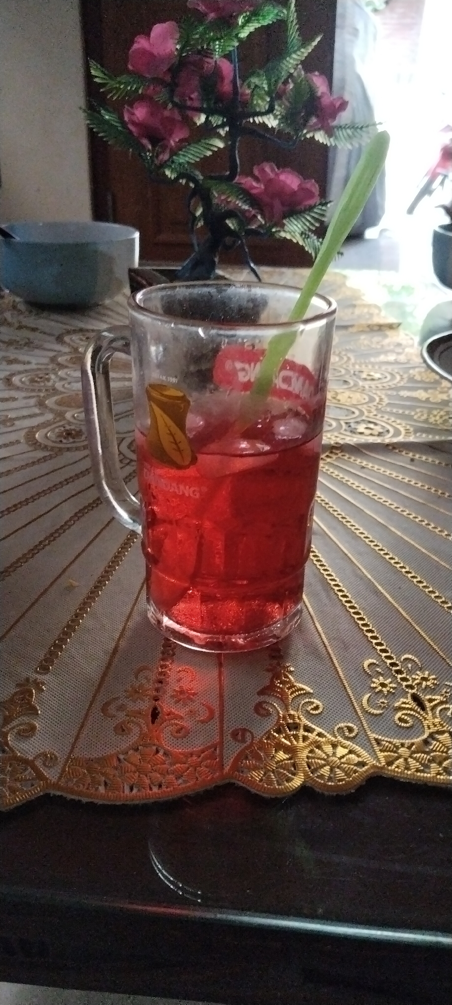 A glass mug filled with a red beverage and ice is placed on a patterned tablecloth. A green straw or herb stem extends from the drink. Behind the mug, a decorative artificial plant with pink flowers is visible. The scene appears to be indoors, with a wooden door in the background. The tablecloth has an intricate design with gold and beige accents.