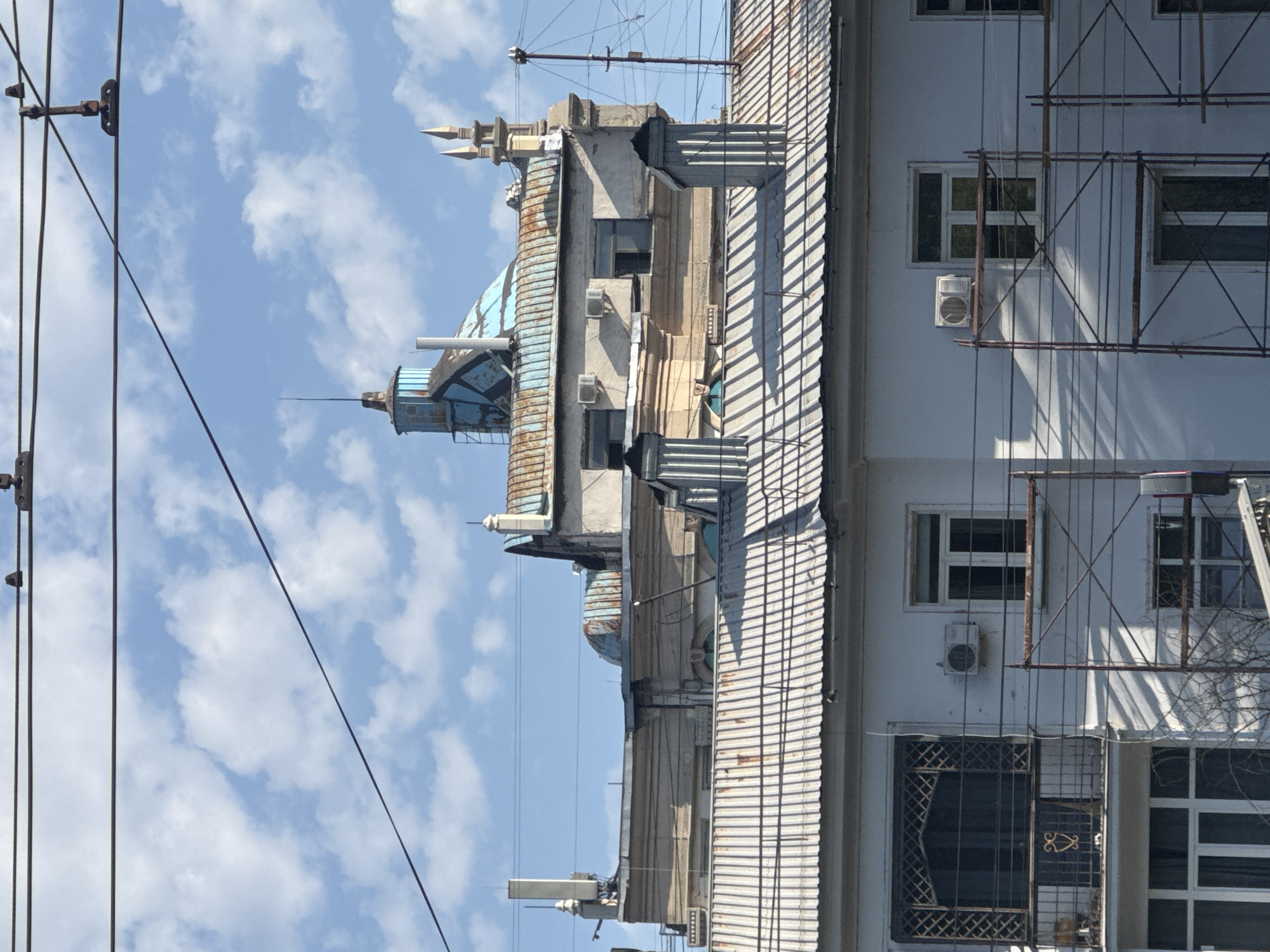 The image shows a view of buildings in Almaty, Kazakhstan, under a sky with scattered clouds. The buildings are multi-story structures with distinct architectural features. The foreground shows a white building with corrugated metal roofing, windows, and a balcony with an ornate metal railing. Scaffolding is erected against this building, suggesting ongoing construction or renovation work.
Behind the foreground building, another structure is visible with a light-colored facade, featuring several windows and air conditioning units. This building has a more ornate architectural style, with decorative moldings and a domed roof section that appears to be covered in a bluish material and is showing signs of rust. There are also two prominent chimneys on the corrugated metal roof in the foreground.
Further in the background, more building structures are visible, including one with a distinctive blue, angular roof with rusted sections and a pointed spire. Another section of the building has multiple small spires or decorative elements at the top. Electrical wires are strung across the scene, indicating the presence of infrastructure within the urban environment. The overall impression is a scene of an aging urban landscape with elements of both residential and older architectural styles.