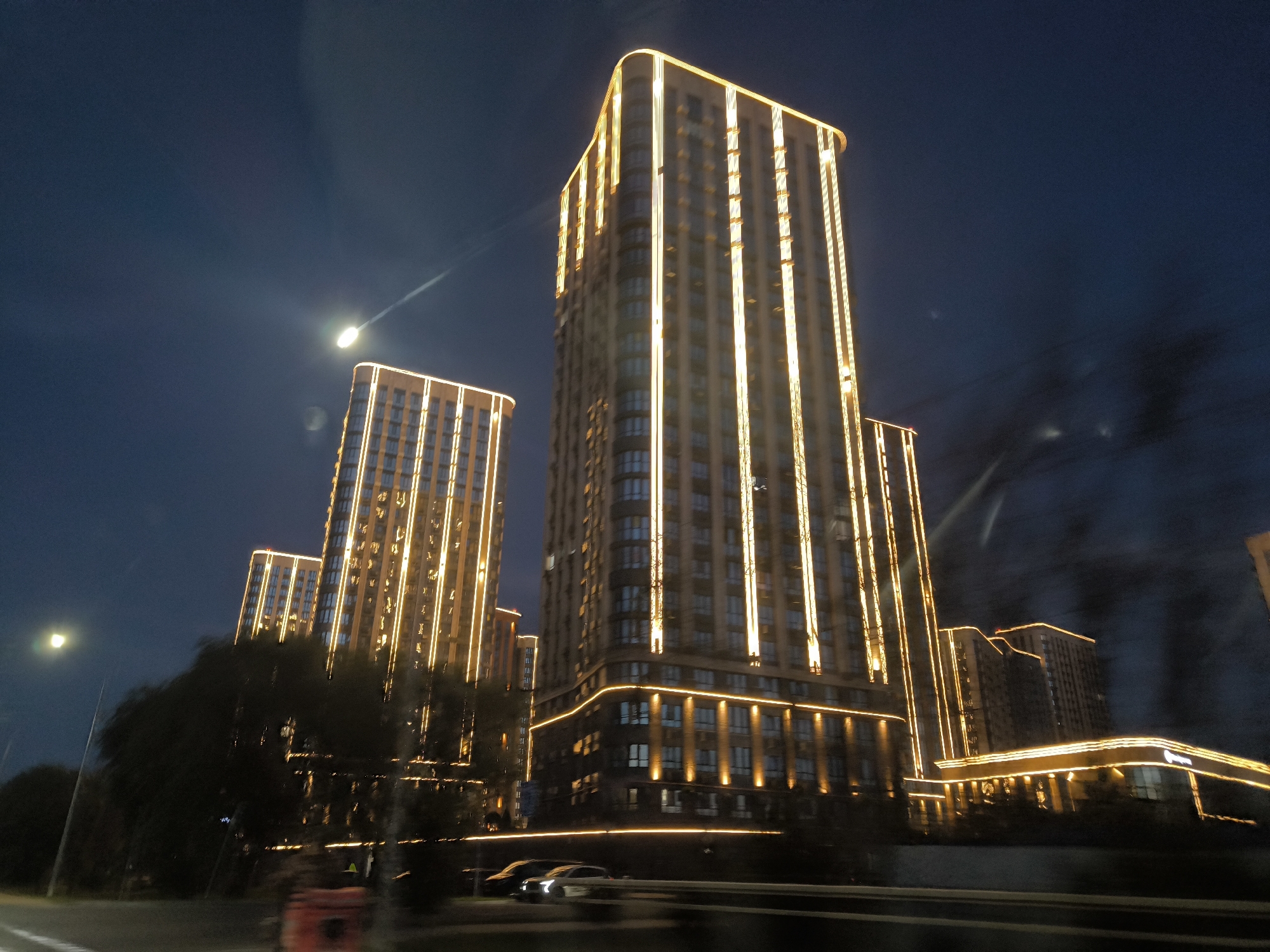 A nighttime exterior shot captures a series of tall, modern buildings illuminated by vertical bands of warm, golden light that outline their facades. The sky is a deep, dark blue, with a faint streak of light and a bright circular orb, possibly the moon or a streetlamp, visible in the upper left quadrant. Several buildings are clustered together, with the tallest in the center dominating the scene. These structures are multi-story residential or office buildings with visible windows. Below the buildings, a dark expanse suggests a road or pathway, with the blurred outlines of a few cars driving by. Trees and greenery are visible in the foreground and between the buildings, adding a natural element to the urban landscape. The overall impression is one of a modern, well-lit city at night.