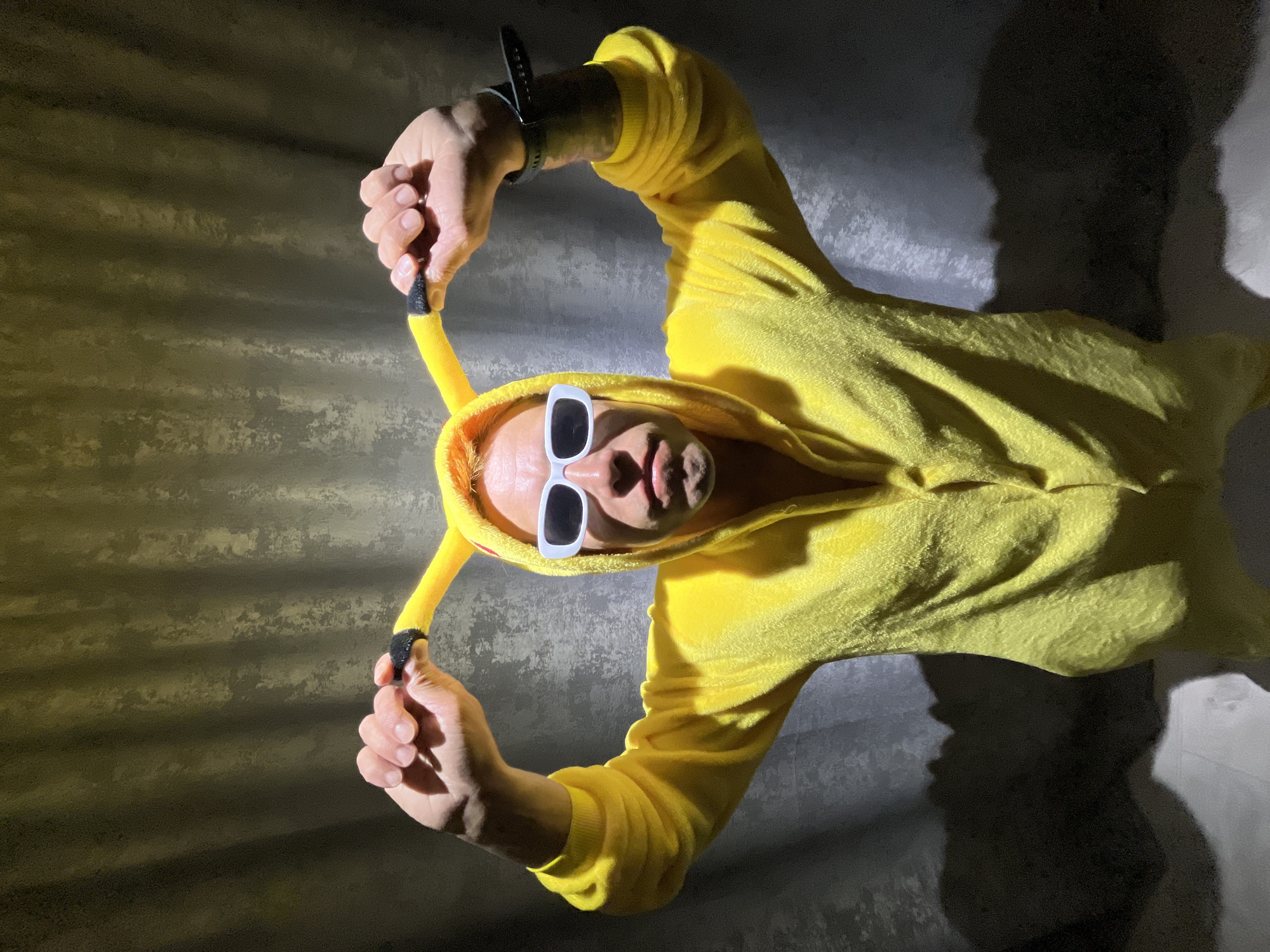 A person is depicted in a full-body yellow suit, likely a kigurumi or onesie, designed to resemble Pikachu with distinct ears tipped in black. The individual wears white, rectangular sunglasses and a dark-colored watch on their left wrist. They are actively holding and pulling the black-tipped drawstrings of the suit's hood with both hands.
The scene takes place indoors, with the person positioned against a textured, light-grey wall that shows horizontal striations and varying shades, suggesting an uneven surface or a specific lighting effect. A strong, angular shadow is cast on the wall to the person's right, indicating a prominent light source from the left, creating a dramatic interplay of light and shadow across the subject and background. The suit itself appears to be made of a soft, fuzzy material, possibly fleece. The person's expression, framed by the sunglasses, is neutral or slightly determined as they look directly towards the camera. There is no visible text in the image. This media file originates from Chaikovsky, Russia.