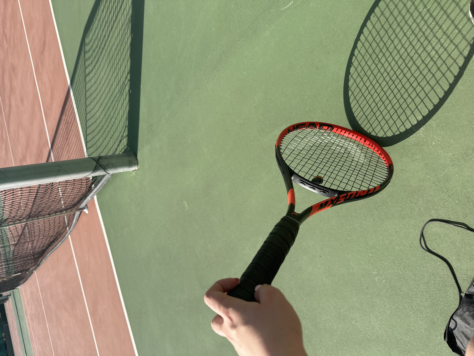 The image shows a person holding a tennis racket on a tennis court. The court is divided into green and brown sections. A net runs across the middle of the court. The racket is a black and red HEAD MXG5 tennis racket, and it is casting a shadow on the court. A tennis ball sits within the strings of the racket. A portion of a bag is visible. The photograph was taken in an outdoor setting. The location is assumed to be Sofia, Bulgaria.