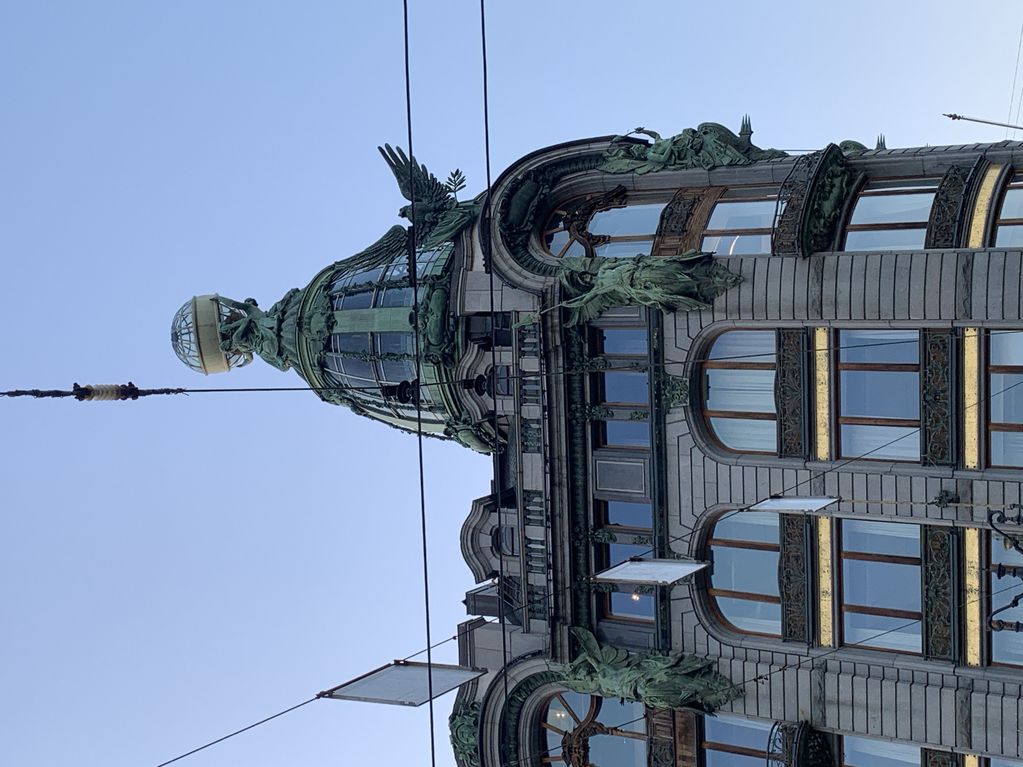 The image shows the Singer House building in Saint Petersburg, Russia, from a low angle looking up. The building's facade is visible, featuring ornate architectural details, including a dome with a glass globe on top, statues, and decorative window frames. A clear blue sky forms the backdrop. Power lines run across the frame, adding to the urban setting.