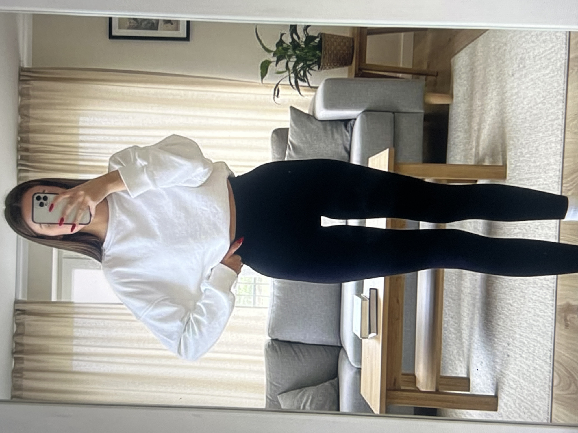A full-body, mirror selfie shows a young woman standing in what appears to be a bedroom or living area. She is wearing a long-sleeved white sweatshirt and black leggings. Her hair is long and dark brown, and she is holding a smartphone in her right hand, taking the picture. The background of the photo reveals a modern living space with a couch, a rug, and large windows with blinds. A potted plant sits on a side table near the couch. The overall impression is casual and relaxed. There is no visible text in the image.