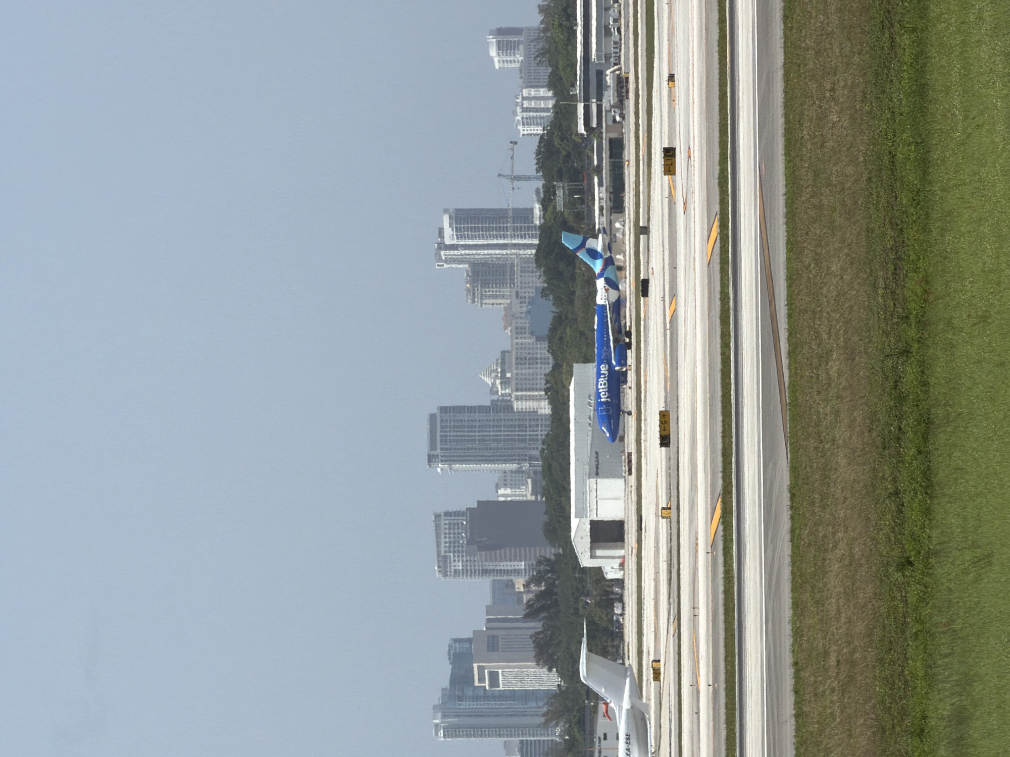 The image depicts an aerial view of an airport in Dania Beach, United States of America, with a city skyline in the background. Two airplanes are visible on the tarmac, one white and one blue with "JetBlue" branding. The airport features multiple runways and taxiways, marked with yellow and white lines. Several airport buildings and structures, including hangars, are also present. The foreground of the image shows a grassy area, likely part of the airport grounds. The sky is clear and pale blue. The overall scene suggests a typical day at a busy airport with ongoing flight operations.