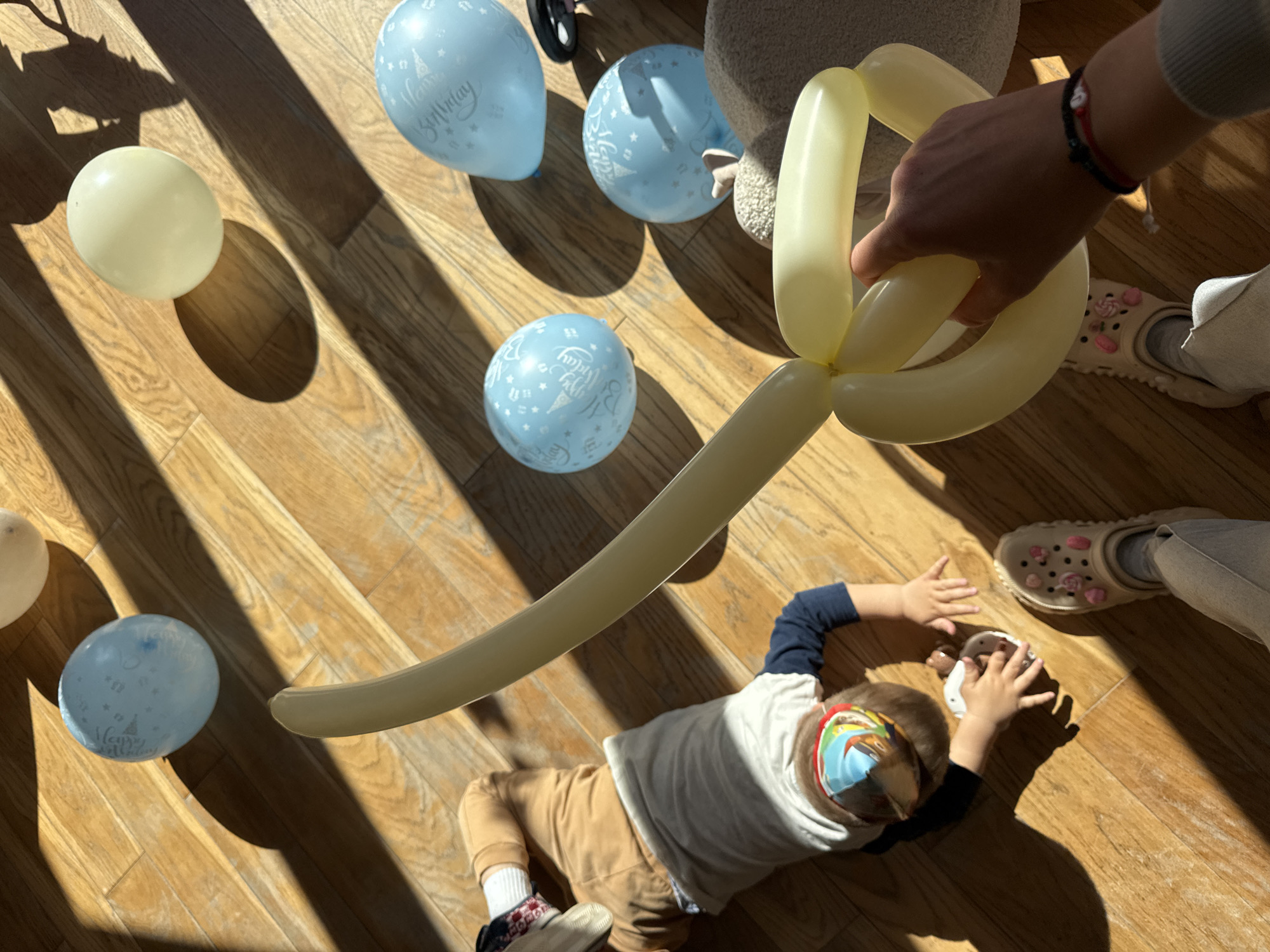 A high-angle shot depicts a child lying on a wooden floor, with several balloons scattered around. The child is wearing a light-colored shirt, brown pants, and patterned socks and shoes. A person's hand is holding a long, yellow balloon animal over the child. There are light blue balloons with "Happy Birthday" written on them. Light-colored balloons are also on the floor. The floor is made of wooden planks, and sunlight is streaming in, creating strong shadows. Part of a person wearing light-colored pants and beige Crocs with decorative embellishments is visible near the child. The scene appears to be indoors, possibly in a private residence. The location in the city of Sofia, Bulgaria, cannot be determined from the information available in the picture.