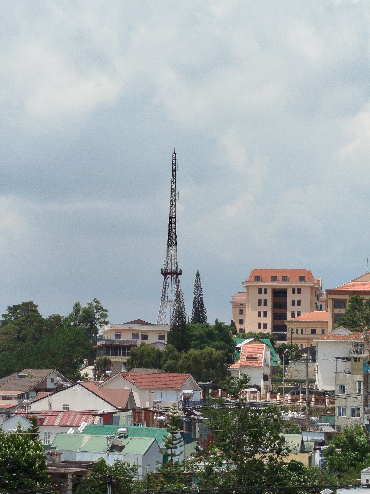 The image depicts a cityscape in Cam Ly - Đà Lạt, Vietnam. The view is elevated, overlooking a cluster of buildings of varying sizes and architectural styles. Predominantly, the buildings are a mix of residential and commercial structures with tiled roofs. The color palette is dominated by earthy tones, whites, and greens, reflecting the natural and built environment. In the middle ground, a tall, lattice-style telecommunications tower dominates the skyline. Patches of trees and foliage intersperse throughout the built environment, adding to the city's aesthetic. The sky is overcast with diffuse light, suggesting a cloudy day. There are no people visible in the image. The overall composition captures the density of the city and the integration of urban development with natural landscapes.