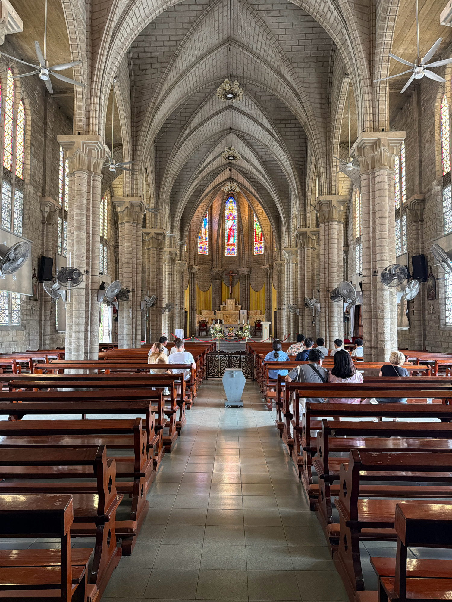The image shows the interior of a large, ornate church, possibly a cathedral, with high vaulted ceilings supported by stone columns. Rows of dark wooden pews fill the nave. Several people are seated in the pews, facing the altar area at the far end of the church. The altar is adorned with religious iconography and stained-glass windows. Multiple large ceiling fans are visible, indicating a warm climate. The overall architectural style suggests a Gothic Revival design. This church is the Nha Trang Cathedral, located in Nha Trang, Vietnam.