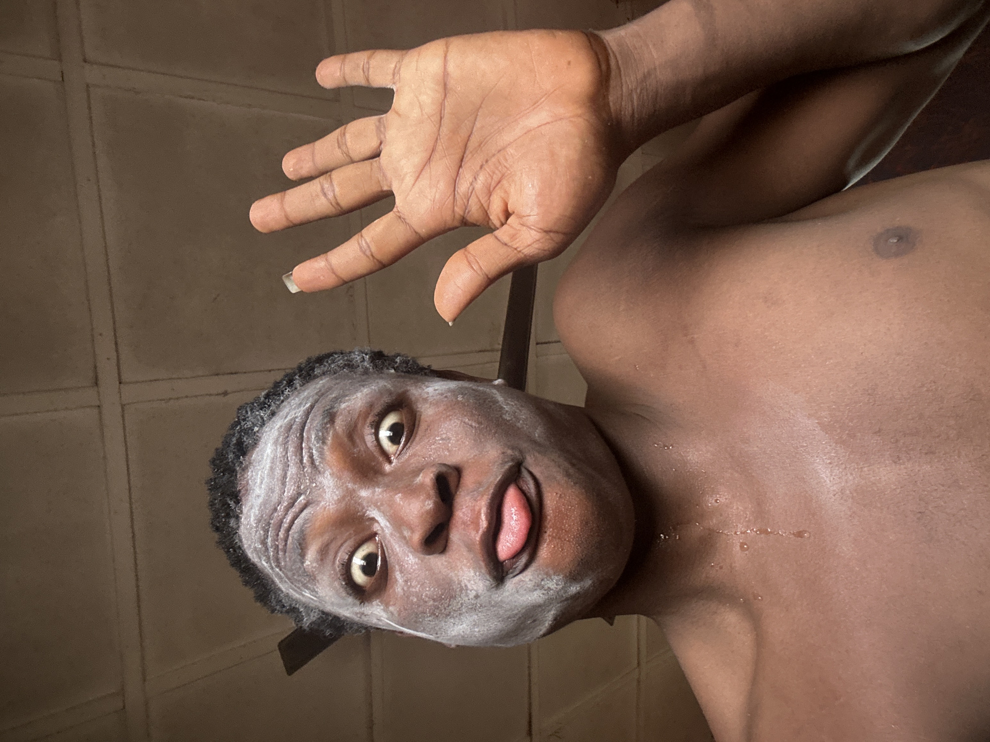 A brown-skinned person is shown from a low angle, face covered in a white substance that appears soapy. The person is sticking their tongue out. The person is topless and has drops of water on their upper chest. The person is holding their right hand up in front of them. The background is a tiled wall. The location is Unknown, Unknown.