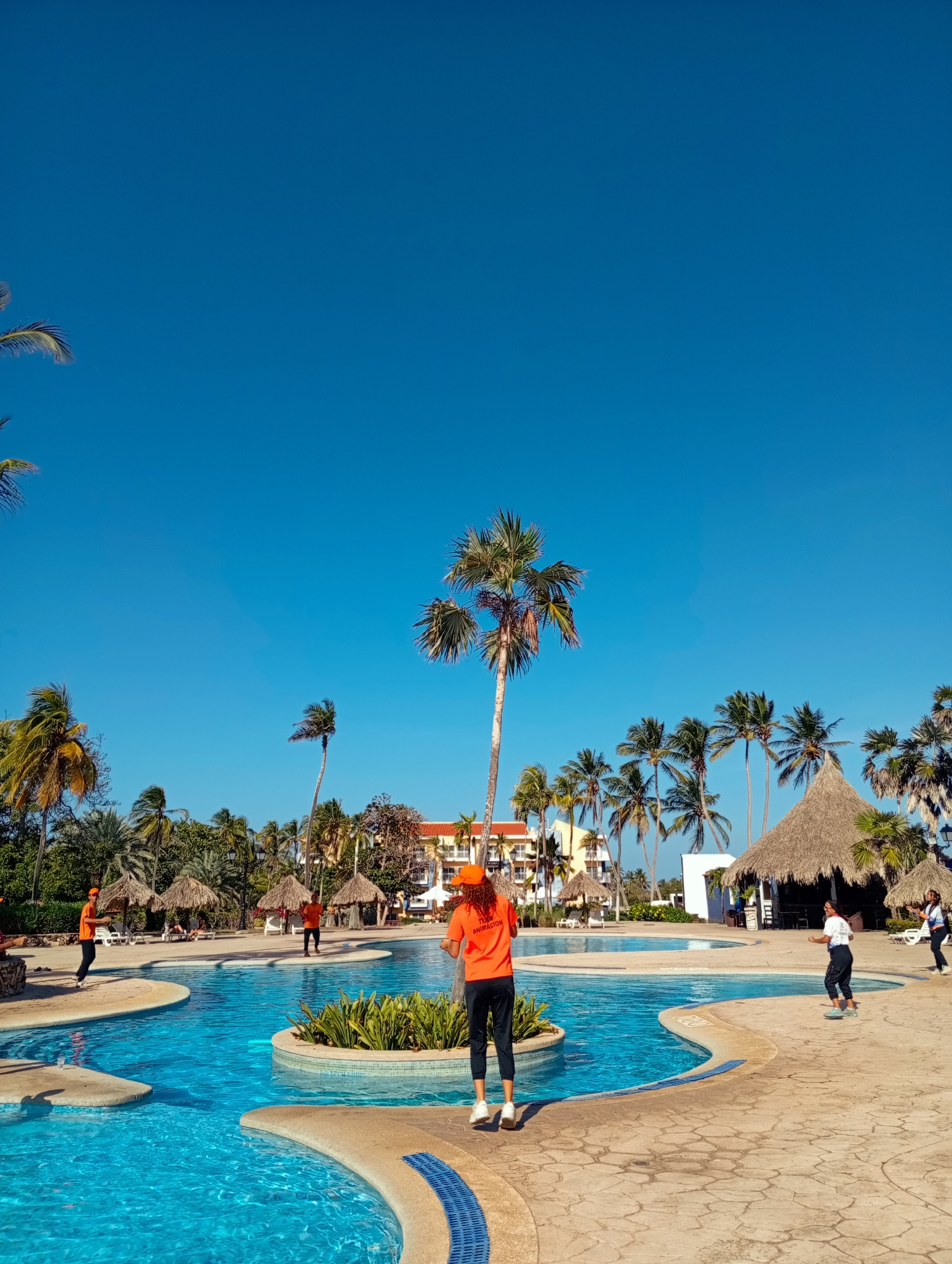The image shows a swimming pool area in Playa El Agua, Venezuela, under a clear blue sky. Several palm trees surround the pool. Four people wearing orange shirts and black pants are positioned around the pool, while two individuals in white shirts and dark pants are also present. The pool is surrounded by paved walkways and several thatched-roof structures, possibly bars or seating areas. A multi-story building is visible in the background behind the palm trees.
