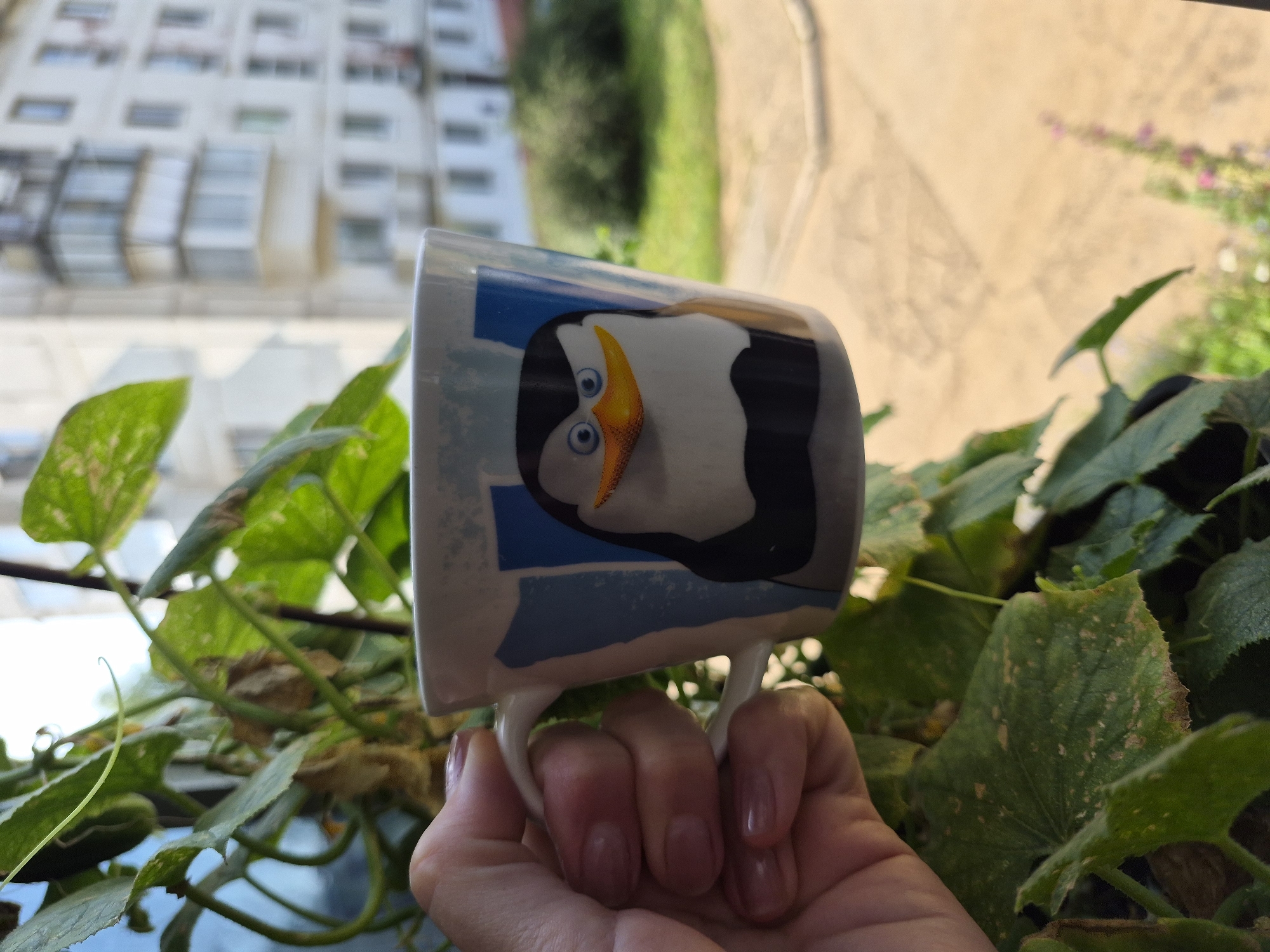 A person's hand, with manicured nails, holds a white ceramic mug decorated with a cartoon penguin against a backdrop of green foliage and a blurry urban environment. The mug features a stylized image of a penguin character with blue eyes and an orange beak against a background of blue and white stripes. In the background, a multi-story building with balconies is visible, characteristic of the architecture found in Chaikovsky, Russia. The lighting suggests it is daytime, with sunlight casting shadows. The overall scene appears to be an outdoor setting, possibly a balcony or window overlooking a residential area.