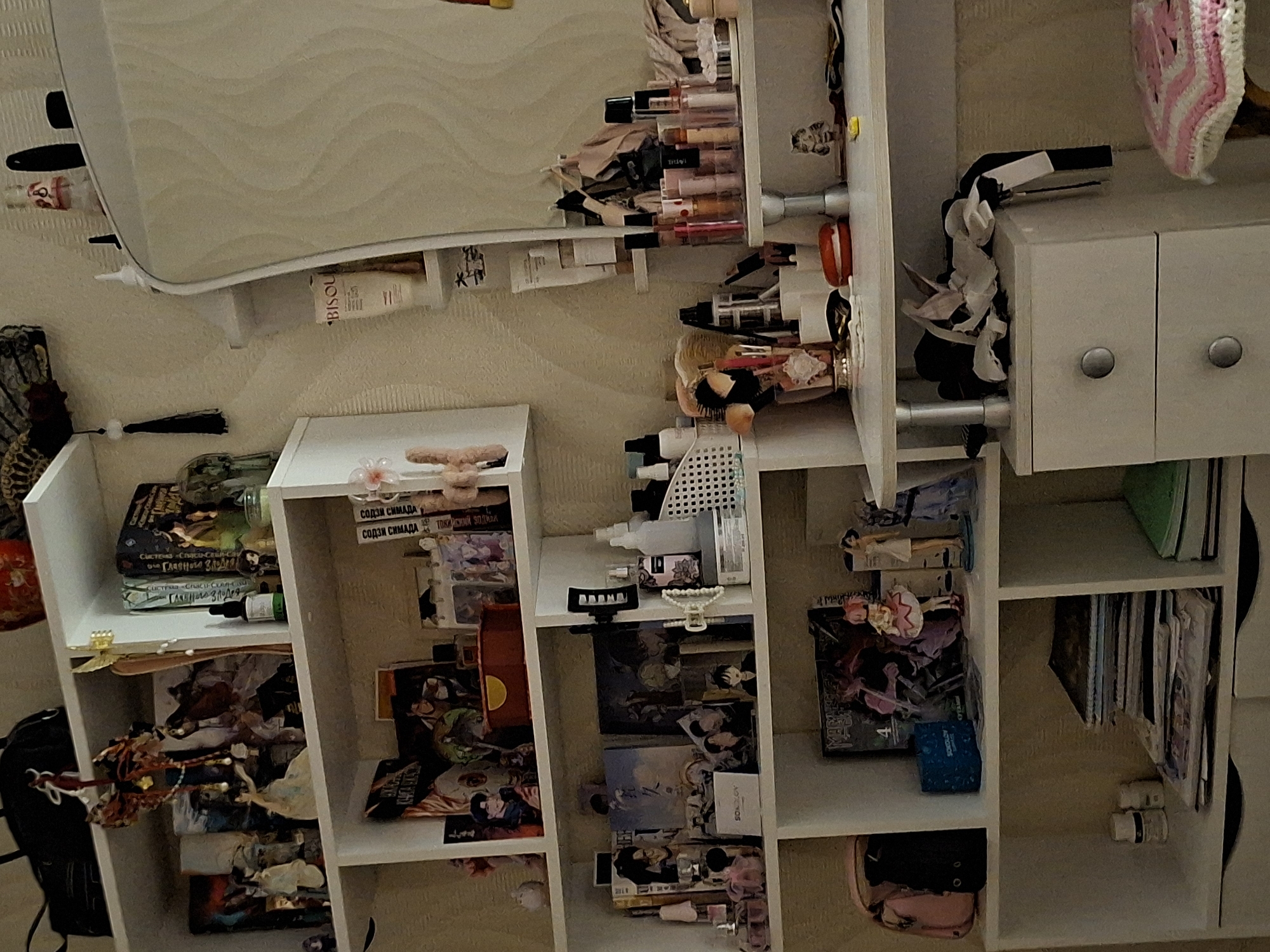 The image displays a densely packed multi-shelf unit and an adjacent vanity area, likely situated in a bedroom or dressing room in Chaikovsky, Russia. The scene appears inverted, with items placed on shelves that would typically be oriented upright.
The primary subject is a white shelving unit, overflowing with personal items. On what appears to be the top surface, a mirror reflects a light-colored wall with a wavy textured pattern. Numerous makeup and skincare products, including bottles and tubes, are arranged here; visible brand names include "BISOU" and "Carme."
To the right, a small white cabinet with two silver-knobbed doors sits on a shelf, with a black stapler and crumpled paper or fabric next to it. A pink and white crocheted piece is also visible.
The shelves below are filled with an eclectic assortment of objects. Several books, some with Russian titles such as "СОДЗИ СИМАДА" (Soji Shimada) and "Главного злодея" (The Main Villain), are stacked. Small figurines, including anime-style characters, are placed among the books and other decorative items. Various small boxes, bearing text like "SOKOLOV," "MONCINOS," and "TRIVI," are also present, alongside bottles, a pearl necklace, and other trinkets. A black backpack and a pink bag are visible on the lower shelves.
The scene portrays a highly personalized space, indicative of interests in beauty products, literature, and potentially anime or collecting. The overall impression is one of organized clutter, with no specific activity taking place. The lighting is consistent with an indoor setting.
