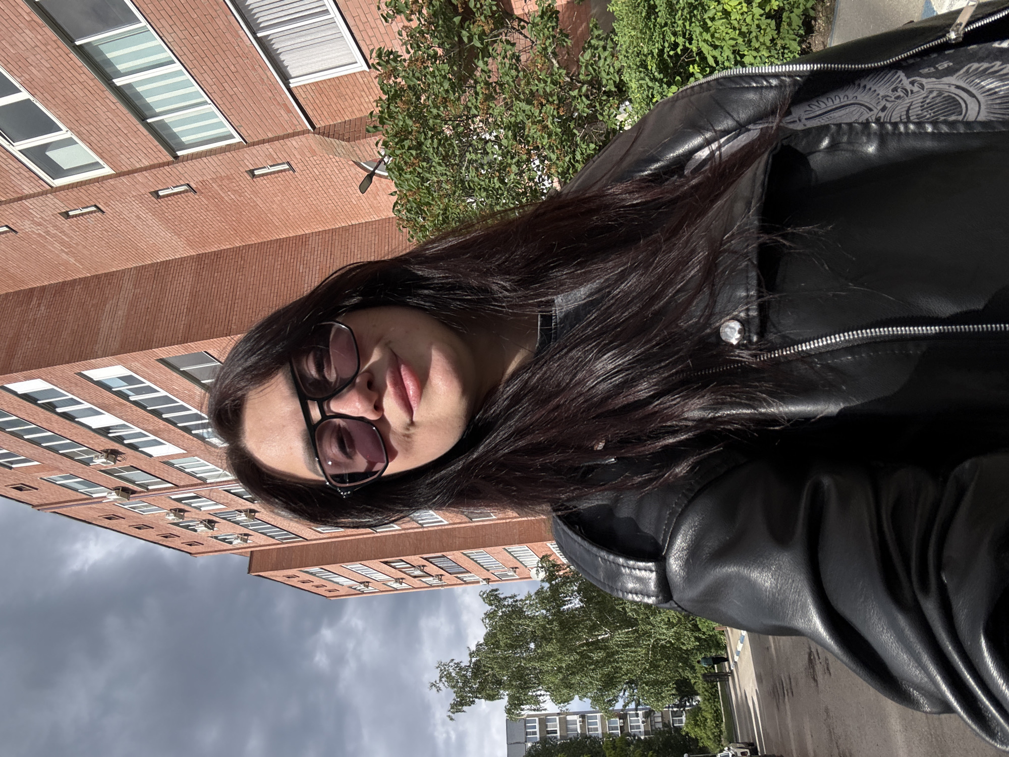 A woman with long, dark hair and glasses is wearing a black leather jacket and standing outdoors. Behind her, there is a large brick building with many windows. Trees are visible, and the sky is overcast. The location appears to be a city.