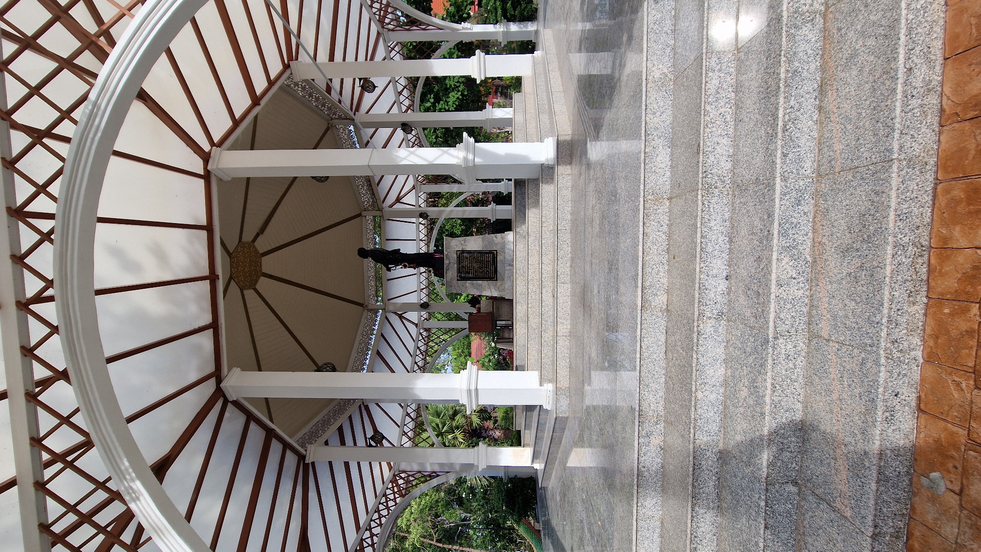 This image depicts a monument situated in the city of Baan Tai, Thailand. The monument is housed within an open-air pavilion with a white, multi-tiered roof supported by numerous white pillars. The pavilion's ceiling features a wooden lattice structure and translucent panels. In the center of the pavilion, on a raised platform reached by granite steps, stands a dark-colored statue atop a stone monument inscribed with text. Surrounding the pavilion are lush green trees and vegetation. The ground in front of the steps is paved with reddish-brown interlocking pavers. The overall scene suggests a memorial or historical site in a tropical setting.