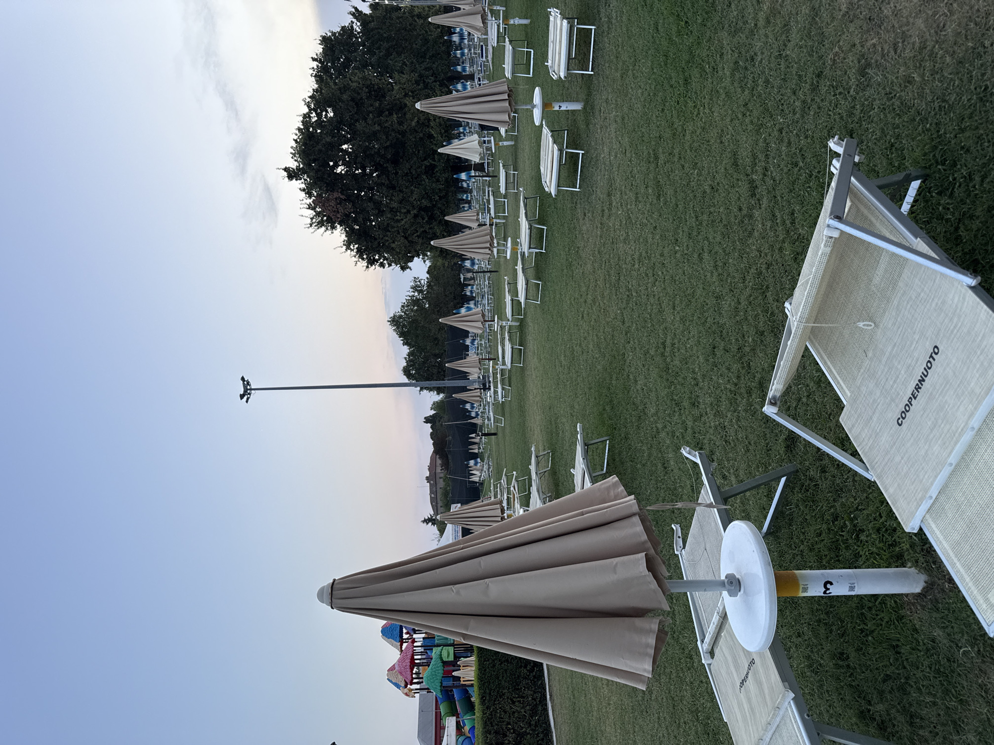 The image depicts an outdoor area, likely a park or recreational facility, with numerous empty lounge chairs and closed umbrellas arranged on a well-maintained lawn. In the background, there is a playground with colorful slides and structures, and trees line the perimeter. A tall lamppost is visible on the right side of the image. The sky is clear and pale blue, suggesting either early morning or late evening. The overall impression is one of an empty, serene space, possibly closed for the day or season. The specific location in Parma, Italy, is not directly identifiable from the image itself, but the scene suggests a public or resort-style amenity.