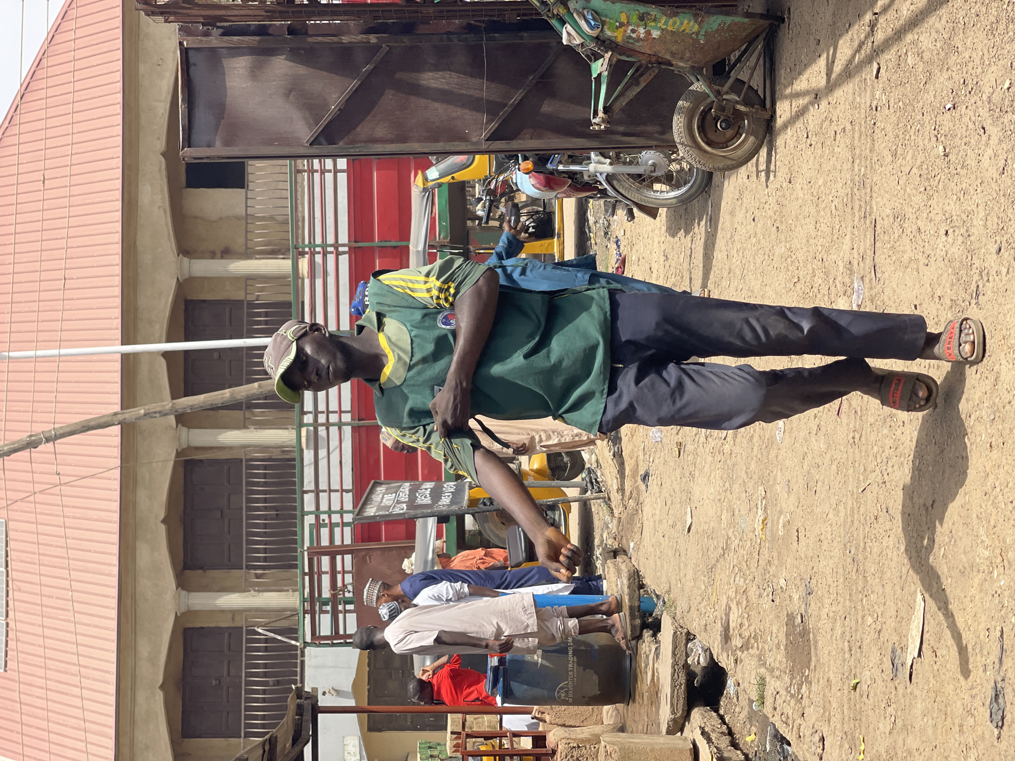 The image depicts a street scene in Zaria, Nigeria. A man wearing a green and yellow shirt, dark pants, a cap, and sandals stands on a concrete sidewalk. He has a tan bag slung across his shoulder. Behind him, several men in traditional Islamic attire, including white robes and caps, are standing and appearing to be using a water station. A three-wheeled motorized vehicle (keke) is parked nearby, as well as another motorcycle. A large metal gate or fence is visible in the background. A sign with writing is displayed near the men. The backdrop includes a building with multiple doors and windows, topped with a corrugated metal roof. The ground is sandy and partially paved. A drain runs along the edge of the sidewalk.