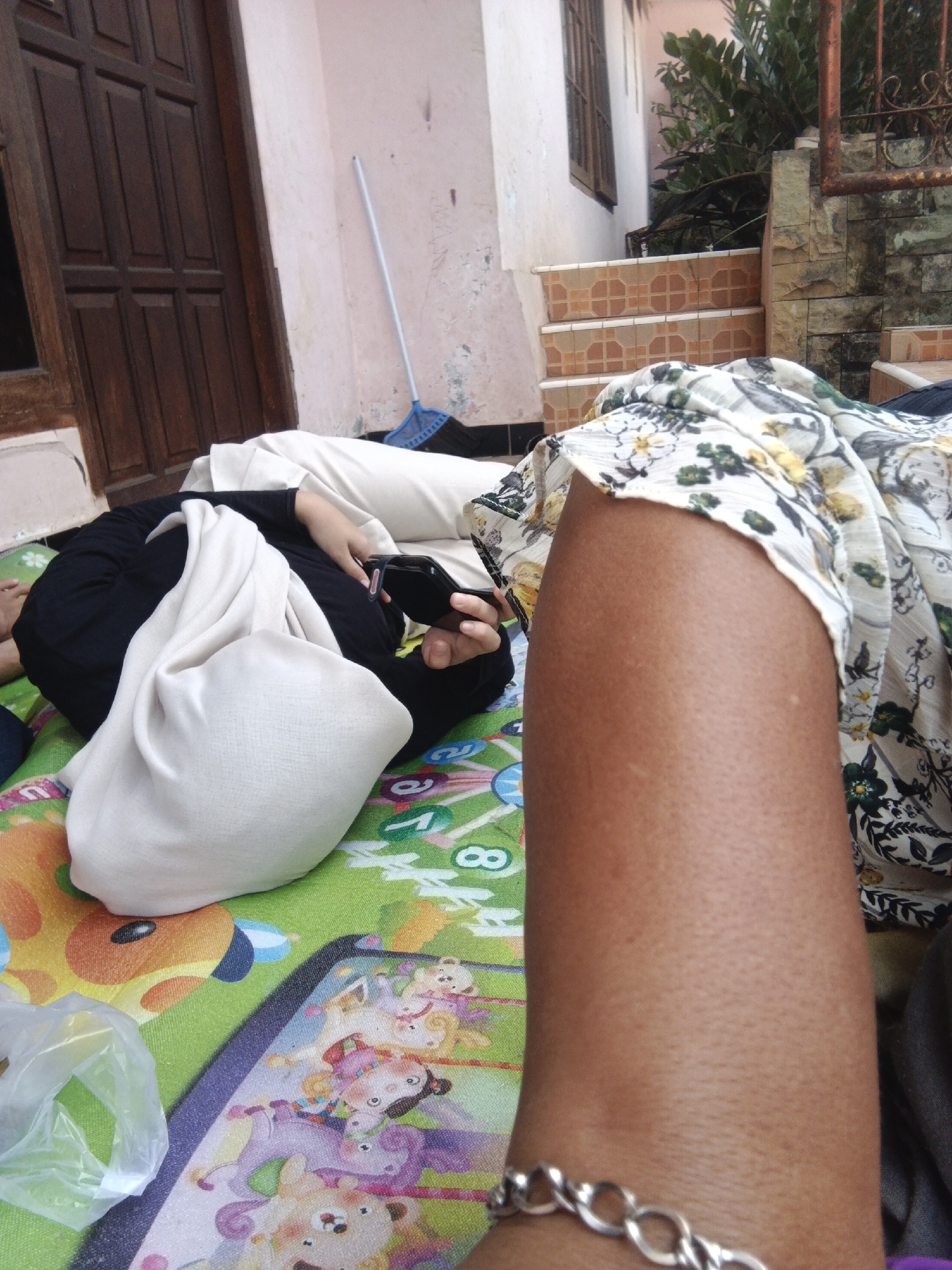 The image depicts a close-up view of two individuals lying down on a colorful mat, likely in an outdoor setting. The mat has cartoonish illustrations of animals and children. One person, on the left, is wearing a black hijab and a white garment. They are holding a black object, possibly a smartphone, in their hand. Their body is partially visible, and they appear to be resting. The other person, on the right, is only partially visible, with their arm and shoulder in focus. They are wearing a floral patterned garment and a silver bracelet on their wrist. In the background, a wooden door and a pink-painted wall are visible. Further in the background, there are stairs with tiled risers, a metal railing, and some greenery, suggesting a residential or public outdoor area. A blue broom with a white handle leans against the wall. The overall scene suggests a casual moment of rest or relaxation. The provided location of Warungkiara, Indonesia, helps to contextualize the environment as a warm climate, consistent with outdoor activities.