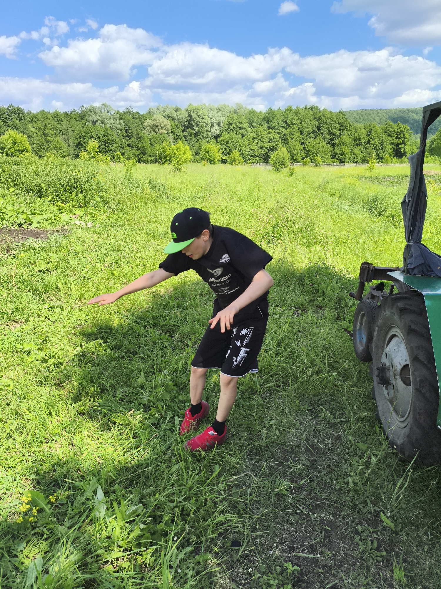 A young male wearing a black t-shirt, black shorts with white trim, a black baseball cap with a green brim, and red sneakers is standing in a grassy field. He is reaching his left hand out towards the grass. To the right of the frame, a portion of a green tractor is visible, including a large wheel and part of its frame. The background consists of a lush green forest under a bright blue sky with scattered white clouds. The scene appears to be in a rural or outdoor setting, possibly in or near the village of Ilmino, Russia.