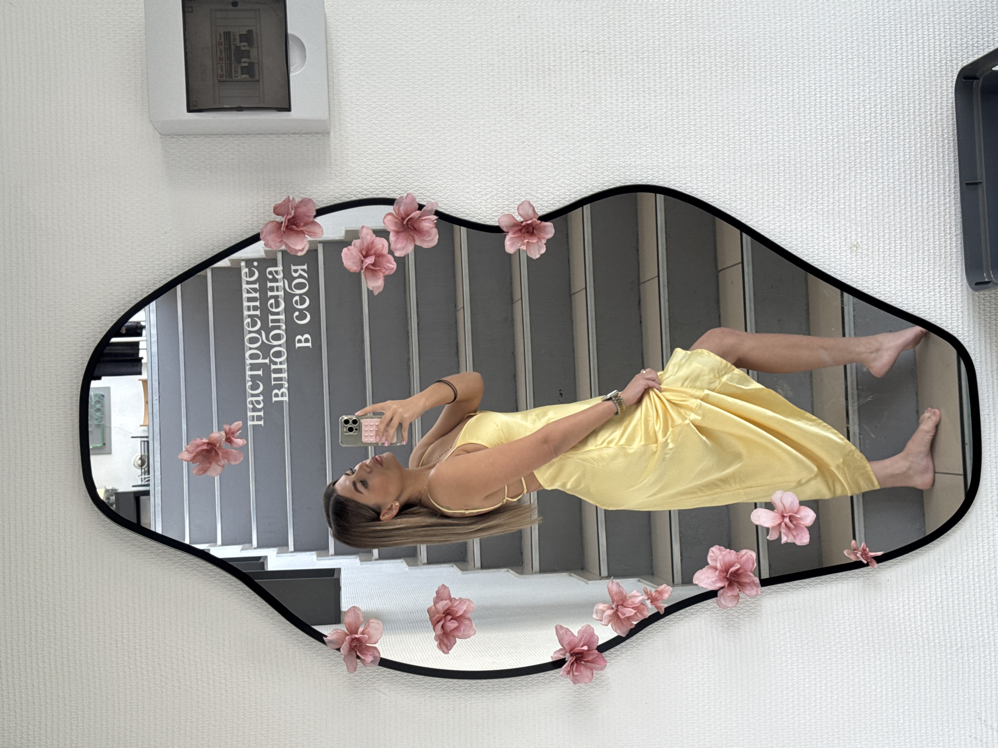 The image is a reflection in a wavy-edged mirror decorated with pink flowers. The reflection shows a woman in a yellow dress taking a selfie on a staircase. A phrase in Russian is visible in the mirror near the top left corner. An electrical panel and another fixture are seen on the wall behind the mirror.
The image does not contain information to verify the location in Chaikovsky, Russia.