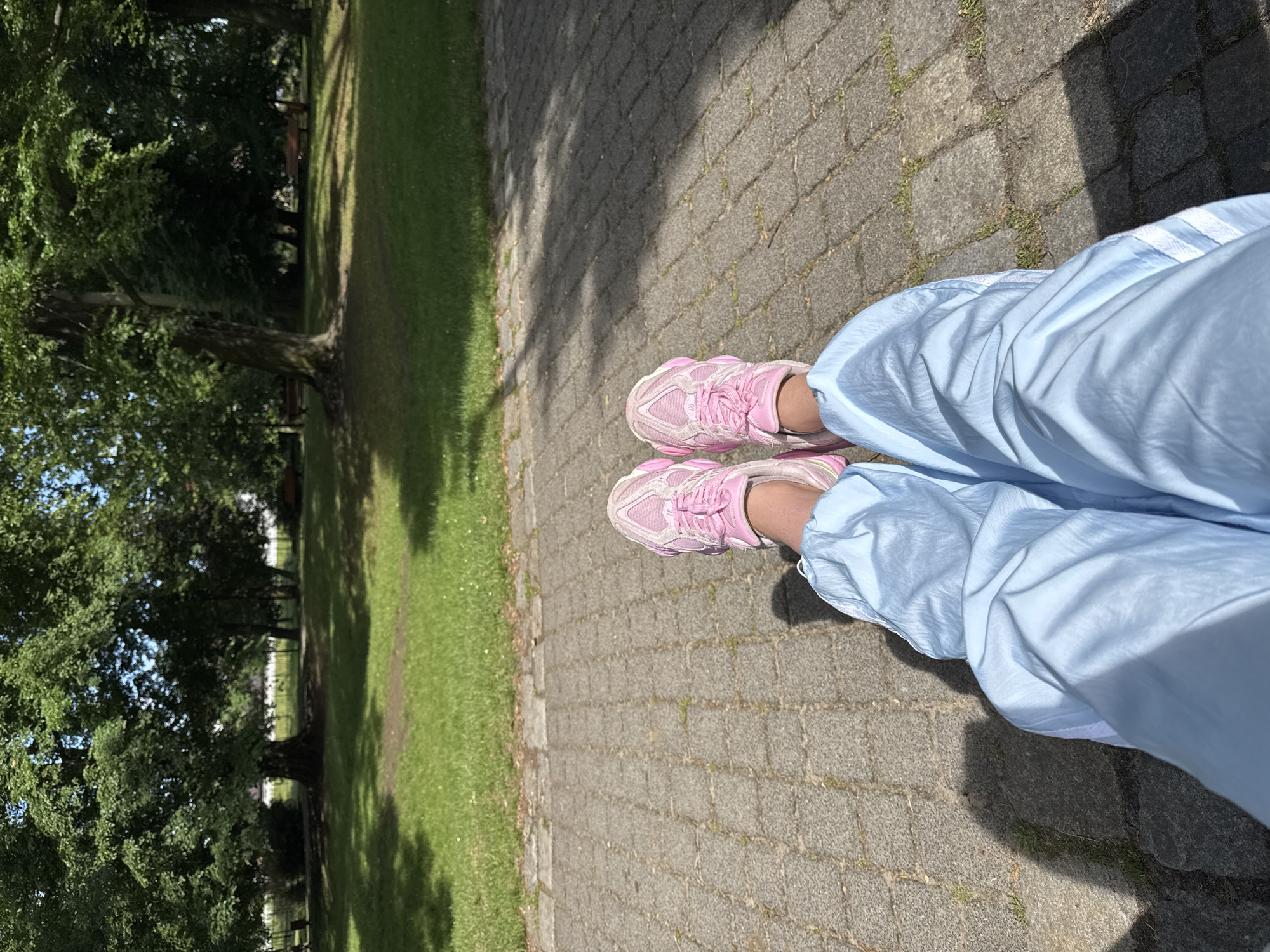 The image displays a first-person, downward-facing view of a person's legs and feet situated on a paved pathway within an outdoor park. The person wears light blue, loose-fitting track pants or joggers with elasticated cuffs and subtle white stripes along the outer seam. On their feet are light pink athletic sneakers featuring a multi-textured design, white laces, and a somewhat chunky sole. Indistinct branding is visible on the side of the shoes.
The pathway is composed of grey, rectangular interlocking pavers, with small patches of grass growing through the cracks. To the left of the path, a wide area of vibrant green grass extends towards a line of mature trees with dense green foliage. These trees cast prominent, elongated shadows across a significant portion of the pathway. In the distant background, partially obscured by the trees, several park benches are faintly visible. The scene is illuminated by bright natural sunlight, suggesting a clear day during the morning or afternoon. The setting is a tranquil public green space. No other individuals are present, and no discernible text is clearly legible on any items.