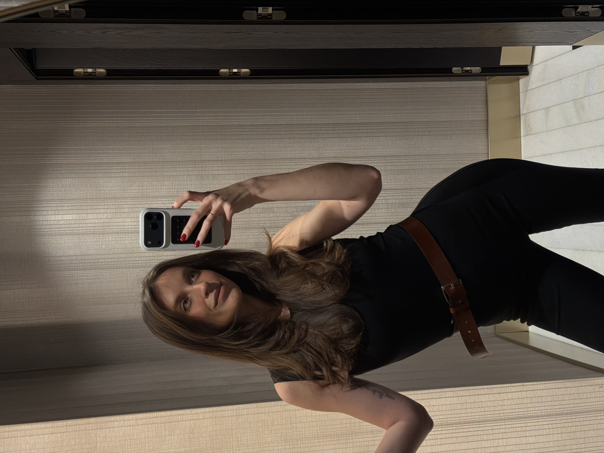 An adult female is depicted taking a mirror selfie in an indoor setting identified as Budva, Montenegro. She is positioned slightly angled, with her gaze directed upwards and to her right. She has long, layered dark brown hair and wears a black sleeveless top or jumpsuit, accessorized with a brown leather belt featuring a metal buckle. Her right hand, adorned with red nail polish, holds a white smartphone with a dark camera module, capturing the image. A tattoo is discernible on her left upper arm. The background consists of a light-toned wall with a subtle vertical striped pattern. To the right, two dark wooden doors with gold-colored accent strips and hinges are visible. The floor is light-colored, possibly marble or tile, bordered by a gold-toned baseboard. The scene is illuminated by artificial light.