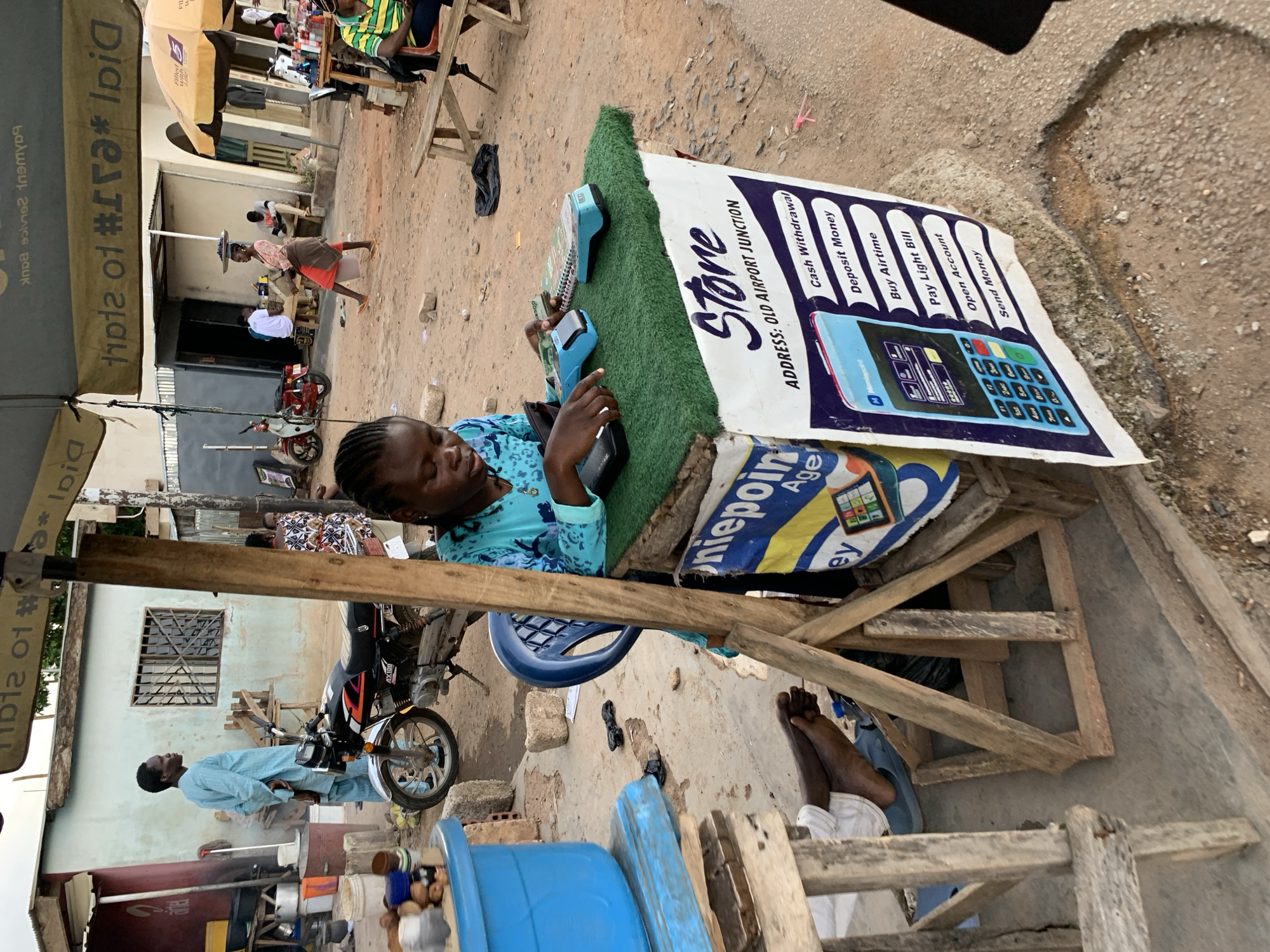 A young woman with braided hair, wearing a blue patterned top, sits behind a makeshift counter covered with green artificial turf. She is operating two point-of-sale terminals and looking at a phone. The counter displays a sign that reads "Store ADDRESS: OLD AIRPORT JUNCTION" and lists services such as "Cash Withdrawal", "Deposit Money", "Buy Airtime", "Pay Light Bill", "Open Account", and "Send Money". Adjacent to the counter, a wooden structure supports a blue plastic basin. In the background, a man in a light blue robe stands next to a motorcycle. The scene appears to be an outdoor market or roadside stall in the city of Jos, Nigeria, with a dusty ground, buildings in the background, and other market stalls visible.