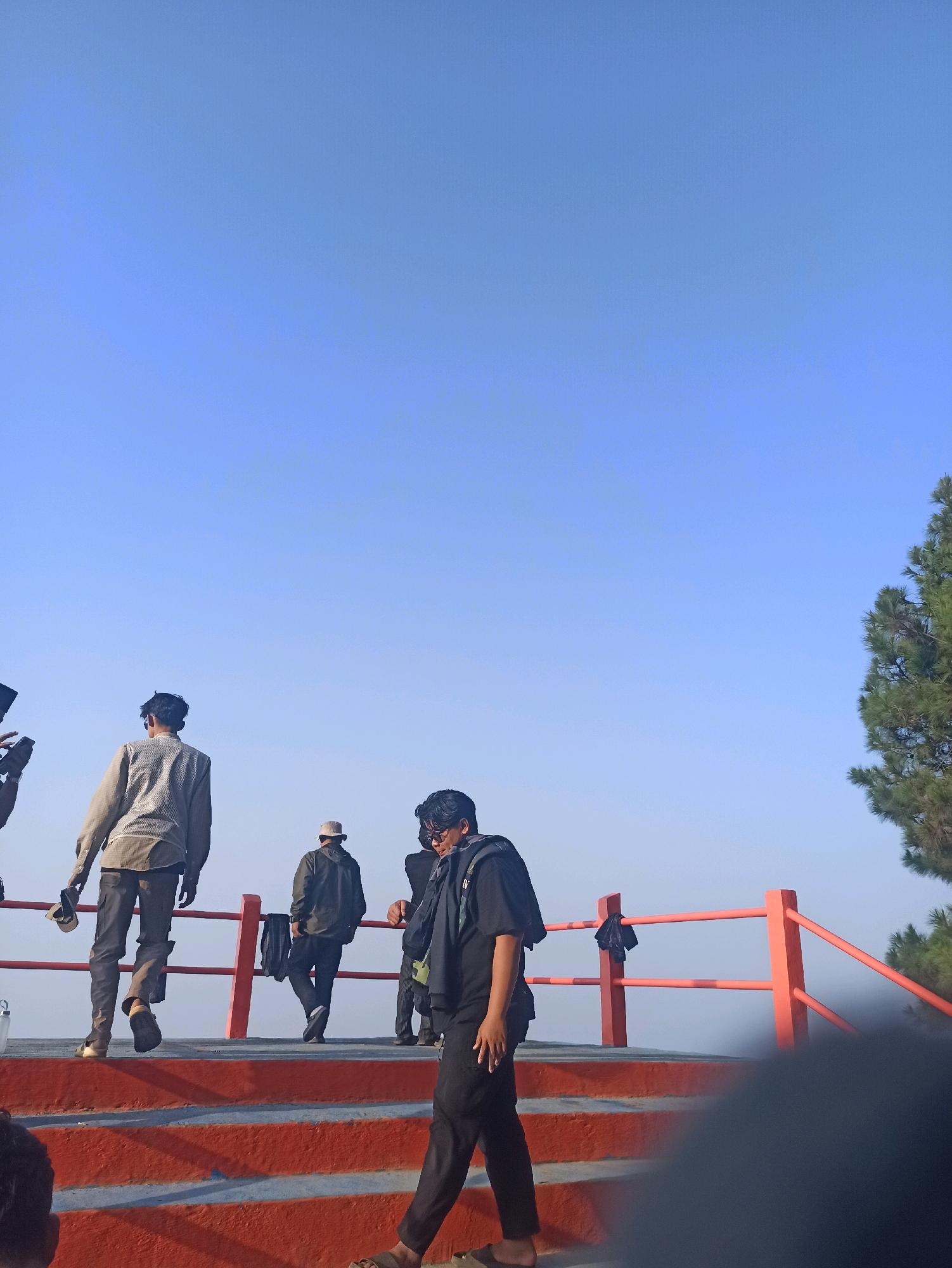 In Cileunyi, Indonesia, a group of five young men are visible on a raised platform with orange stairs and railings, under a clear blue sky. The foreground is dominated by the orange steps and a blurred figure in dark clothing walking down them. To the left, a man in a light-colored, patterned shirt and dark pants walks away from the camera, holding a cap. Further back, a man in a dark jacket and hat walks towards the right. To the right of him, another man in dark clothing is visible. A pine tree stands to the far right of the frame. The men appear to be in casual attire, suggesting a leisure activity or an outing.