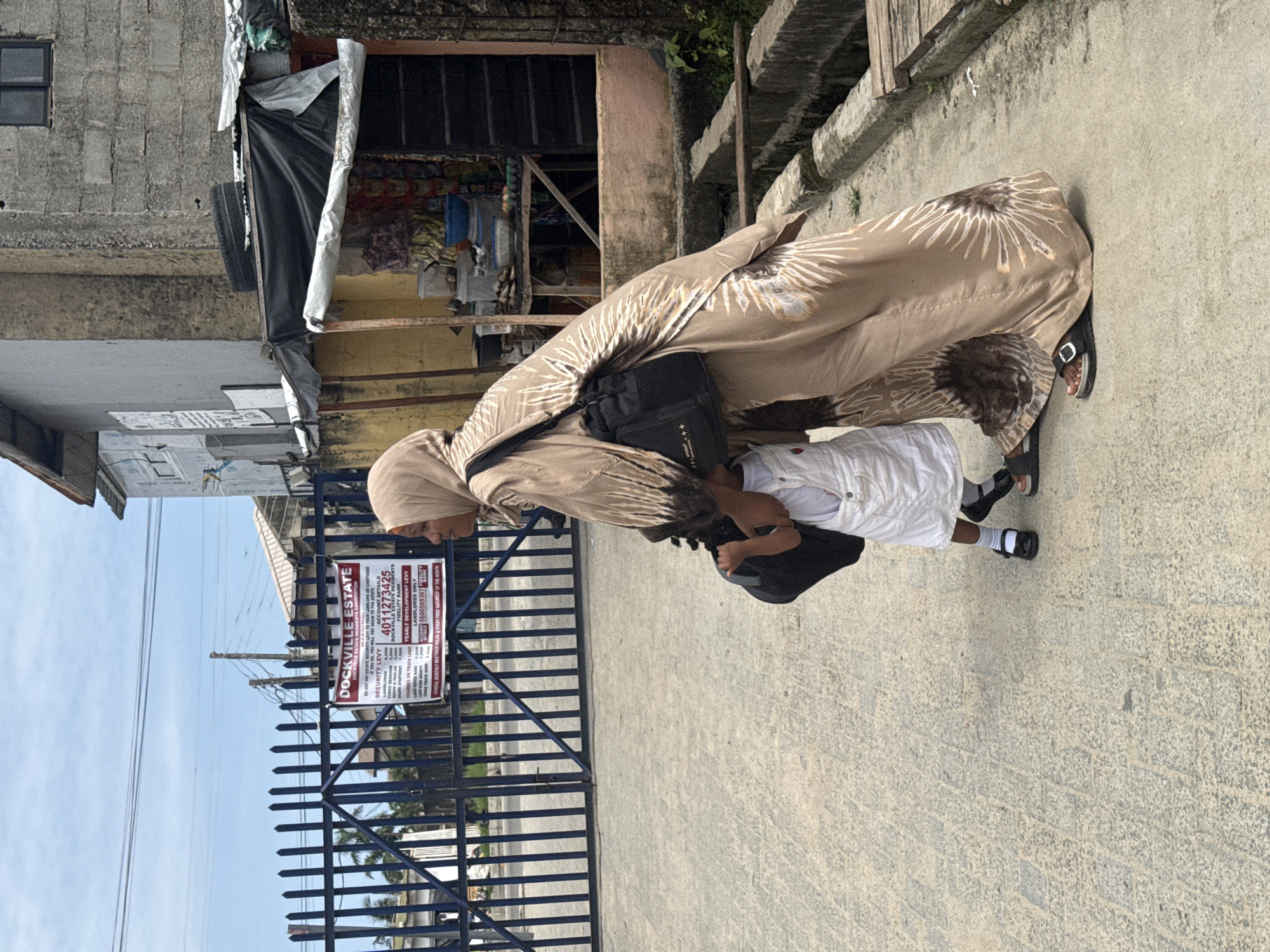 An outdoor, brightly lit image, rotated 90 degrees clockwise, captures a woman and a child walking on a paved surface next to a dark metal gate in Ado, Nigeria. The woman, seen mostly from behind, wears a long, flowing beige garment with a prominent white and brown sunburst pattern, a matching head covering, and black sandals. She carries a black shoulder bag and holds the hand of a young child, who is dressed in white shorts and a white short-sleeved top, also wearing black sandals and white socks.
In the background, beyond the gate, are various structures including concrete buildings, some appearing partially constructed or with makeshift coverings. Power lines are visible against a clear, bright sky.
A prominent sign attached to the gate provides details for "DOCKVILLE ESTATE." The text on the sign includes a "WARNING:" about security levies, stating that if not paid to the landlord or caretaker, residents "WILL PAY AGAIN TO THE ESTATE." It lists "SECURITY LEVY" amounts for different vehicle types (CAR, TRUCK, TRICYCLE, MOTORCYCLE, OTHERS), as well as "YEARLY DEVELOPMENT LEVY" for landlords. Bank account details are provided for "DOCKVILLE ESTATE RESIDENTS" (Fidelity Bank, 4011273425) and "LANDLORDS ONLY" (5600569367). It also states that "GENERAL MONTHLY MEETINGS HOLDS & EVERY FIRST SATURDAY OF THE MONTH." The scene suggests a structured residential community entrance.