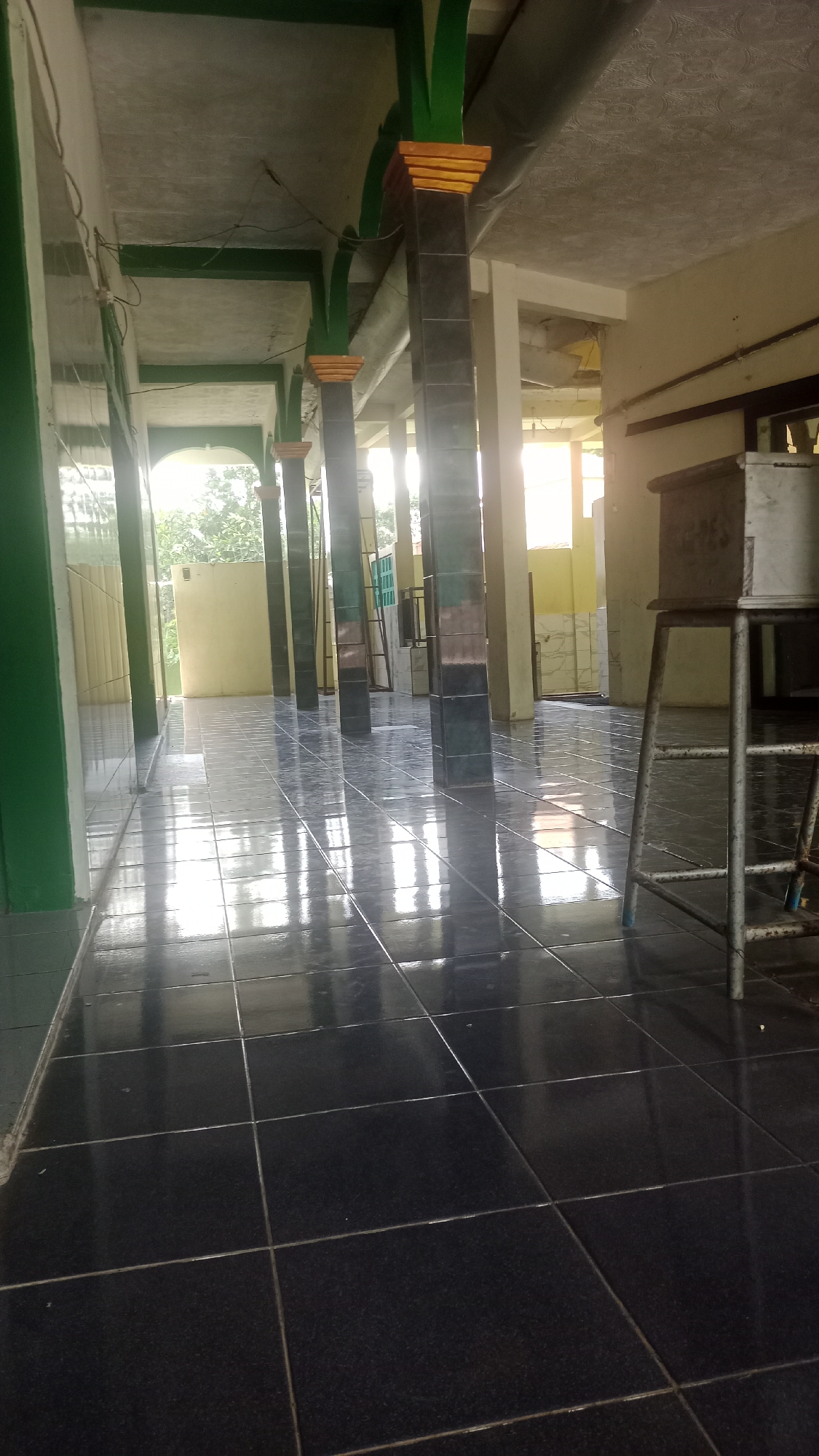 The image depicts a covered walkway with a tiled floor, likely part of a public building or mosque, located in Karyamandala, Indonesia. The walkway is supported by dark, rectangular pillars, each topped with green, orange, and red decorative elements. Sunlight streams in from an opening at the end of the walkway, creating reflections on the glossy dark floor tiles. Green painted walls are visible on the left side, and a yellow wall is seen on the right. In the distance, trees and foliage can be observed through an open doorway. To the right, a metal stand with a rectangular object on top is present. The scene is unoccupied and presents an unobstructed view of the architecture and its surroundings.
