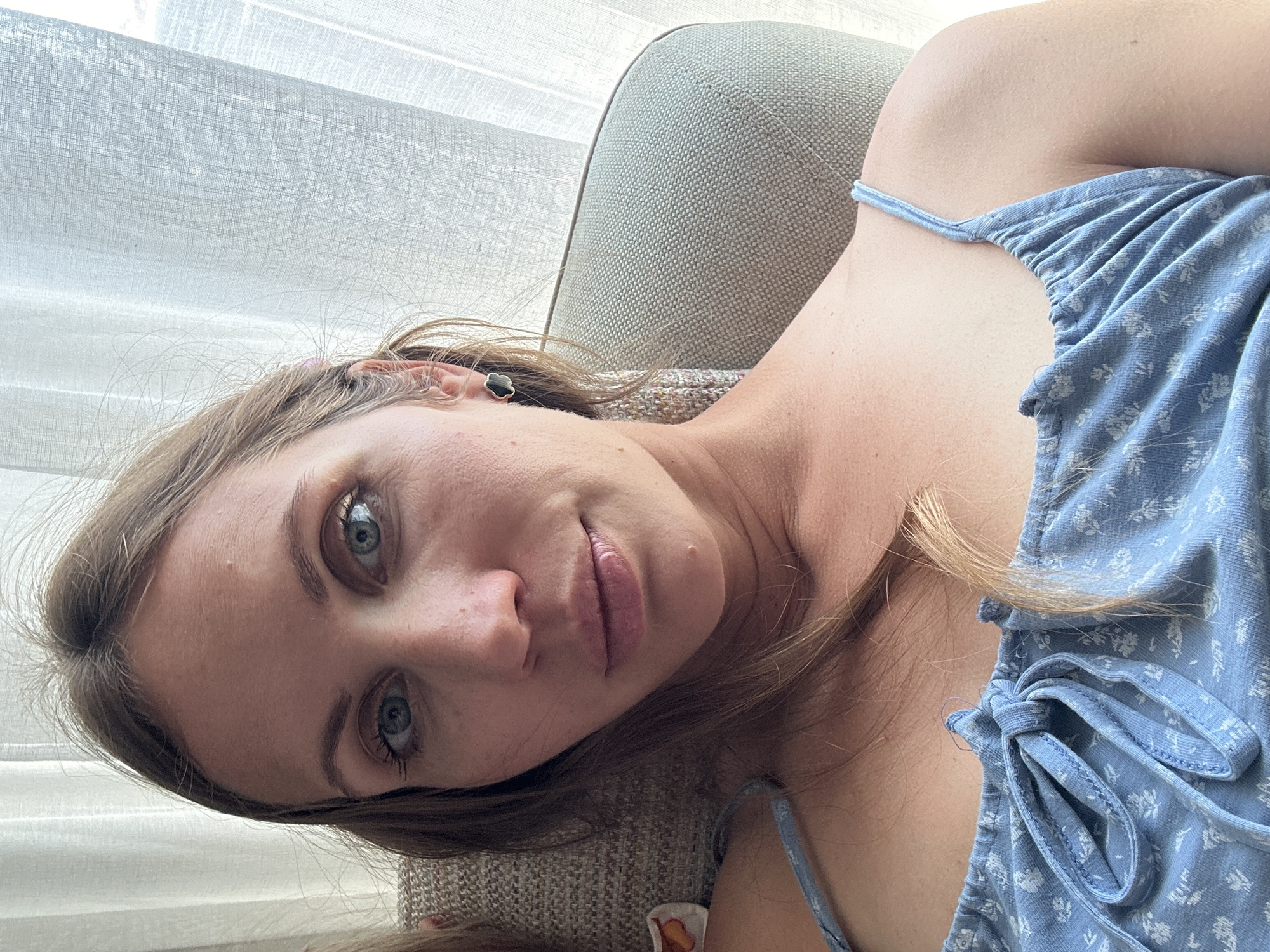 A woman with light brown hair and blue eyes is lying on a light-colored armchair. She is wearing a light blue patterned top. The background shows a light-colored curtain and a glimpse of the armchair. The location is not identifiable as Fethiye, Turkey, based on the visual elements of the image.
