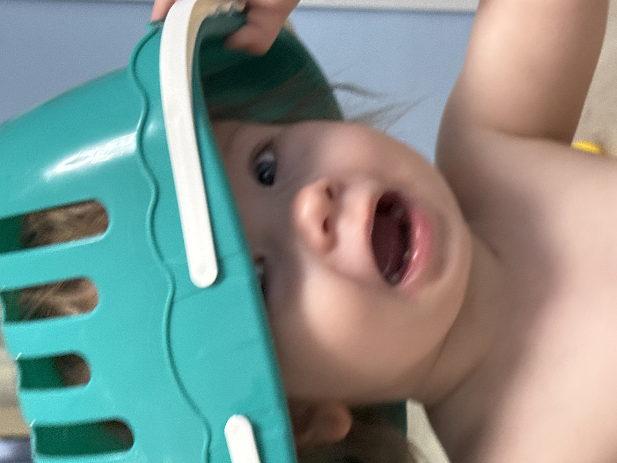 A child is positioned upside down with a teal plastic basket partially covering their head. The child's mouth is open in an apparent expression of surprise or excitement. The child's torso and arm are visible. The background is blurry, showing a light blue surface. There is no indication of the location being Moscow, Russia.